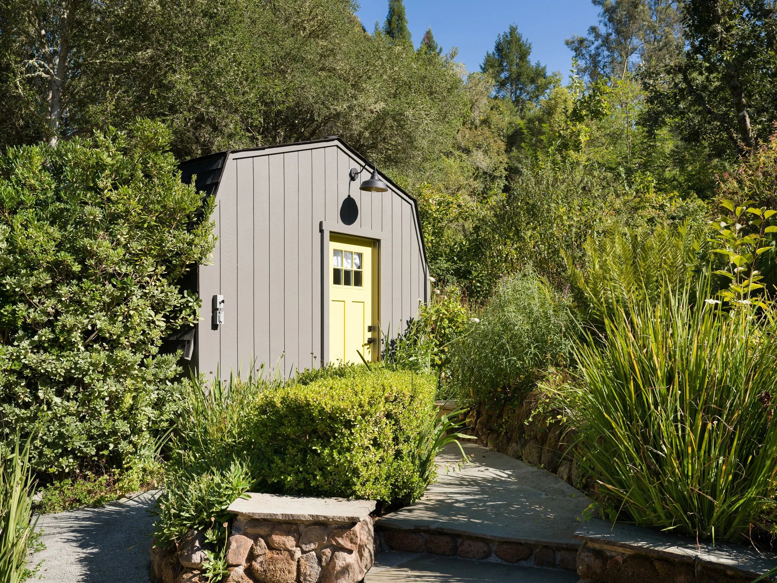 A small gray building with a yellow door and black roof, surrounded by lush green bushes and trees, with a concrete pathway in front and a blue sky above.