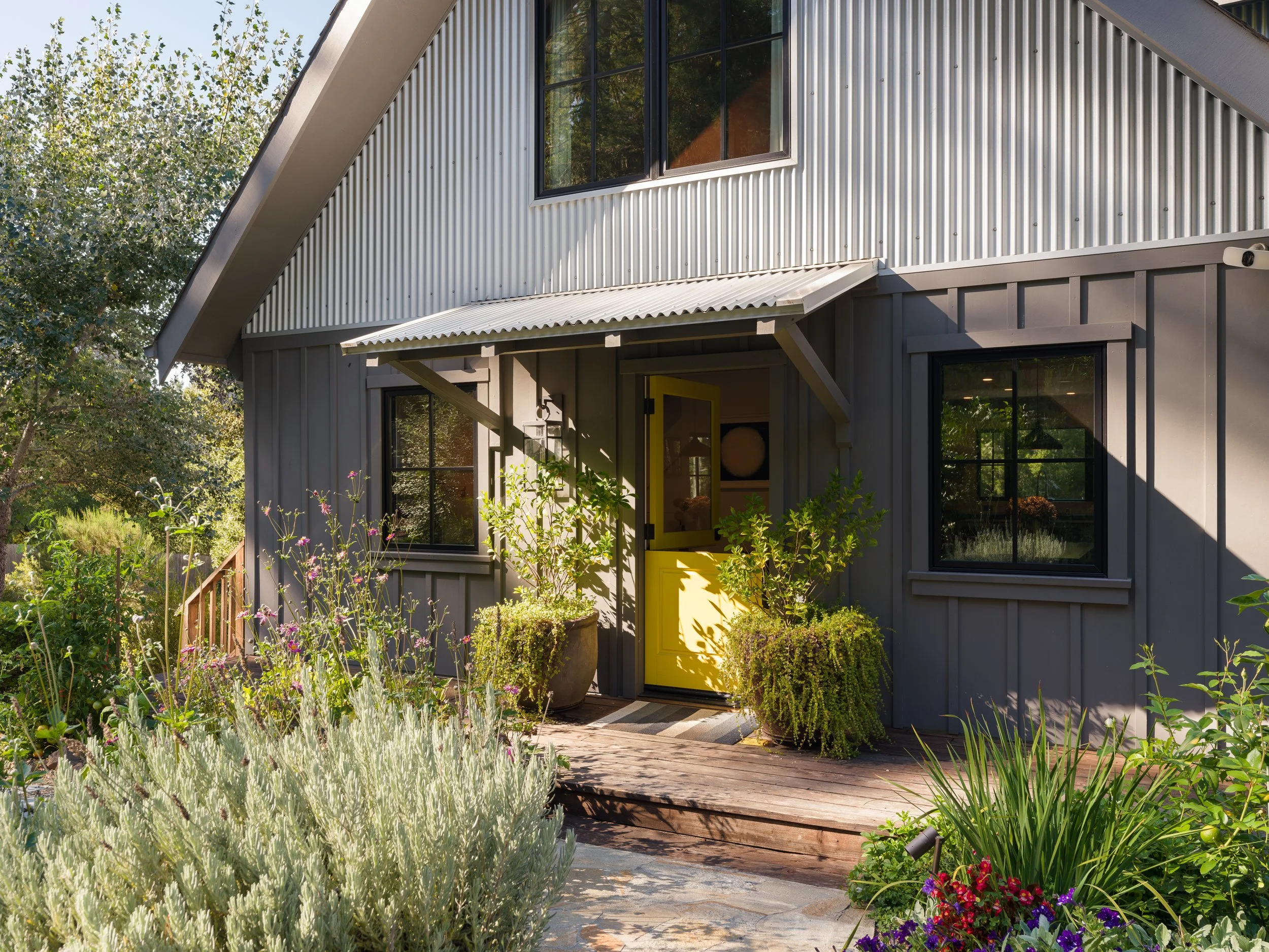 View of a modern house with a yellow door, black window frames, and corrugated metal siding, surrounded by a lush garden with various plants and flowers.