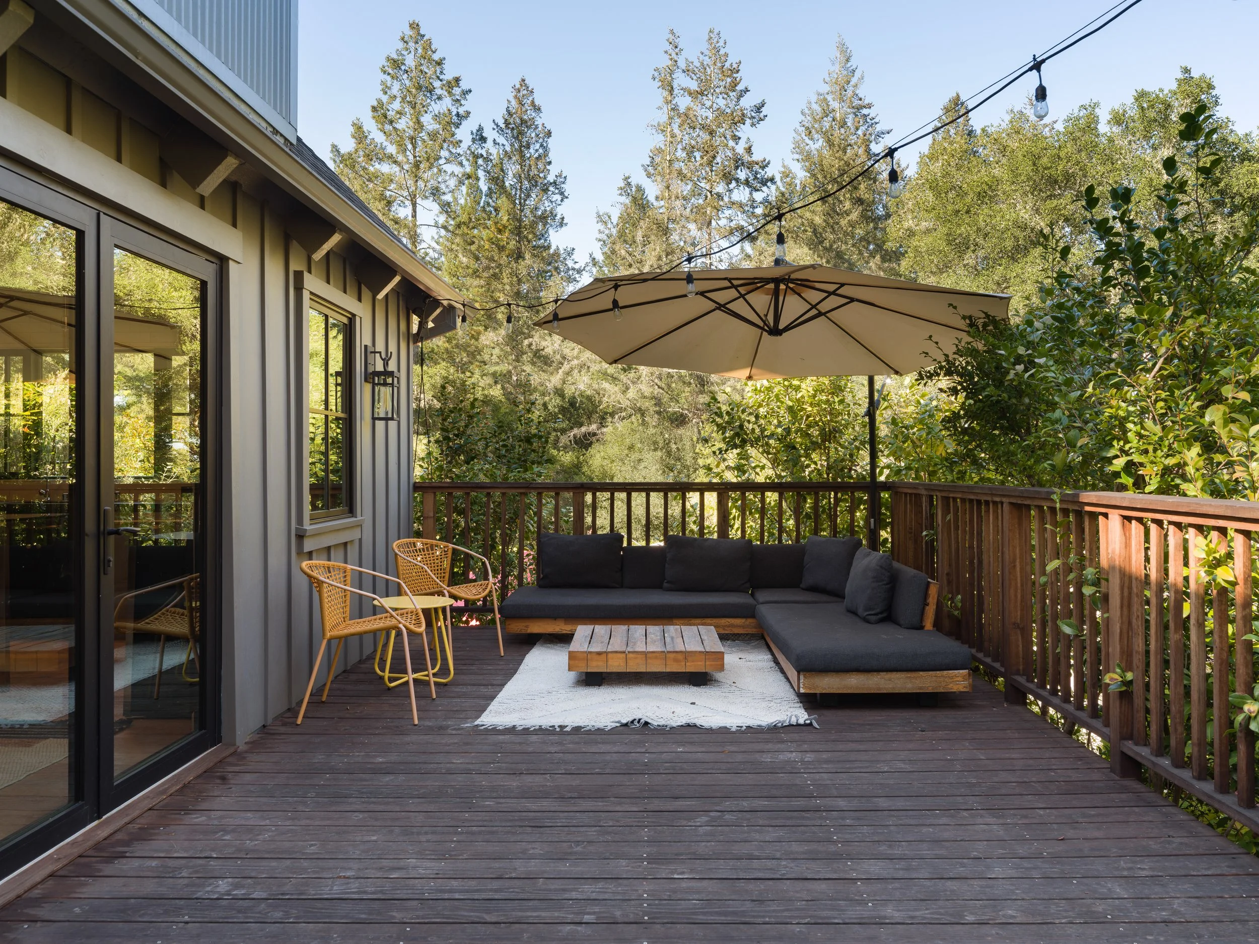 An outdoor wooden deck with a seating area that includes a black outdoor sofa, a wooden coffee table, and some yellow wicker chairs. There is a large beige patio umbrella and string lights overhead. The deck is surrounded by lush greenery and tall tr