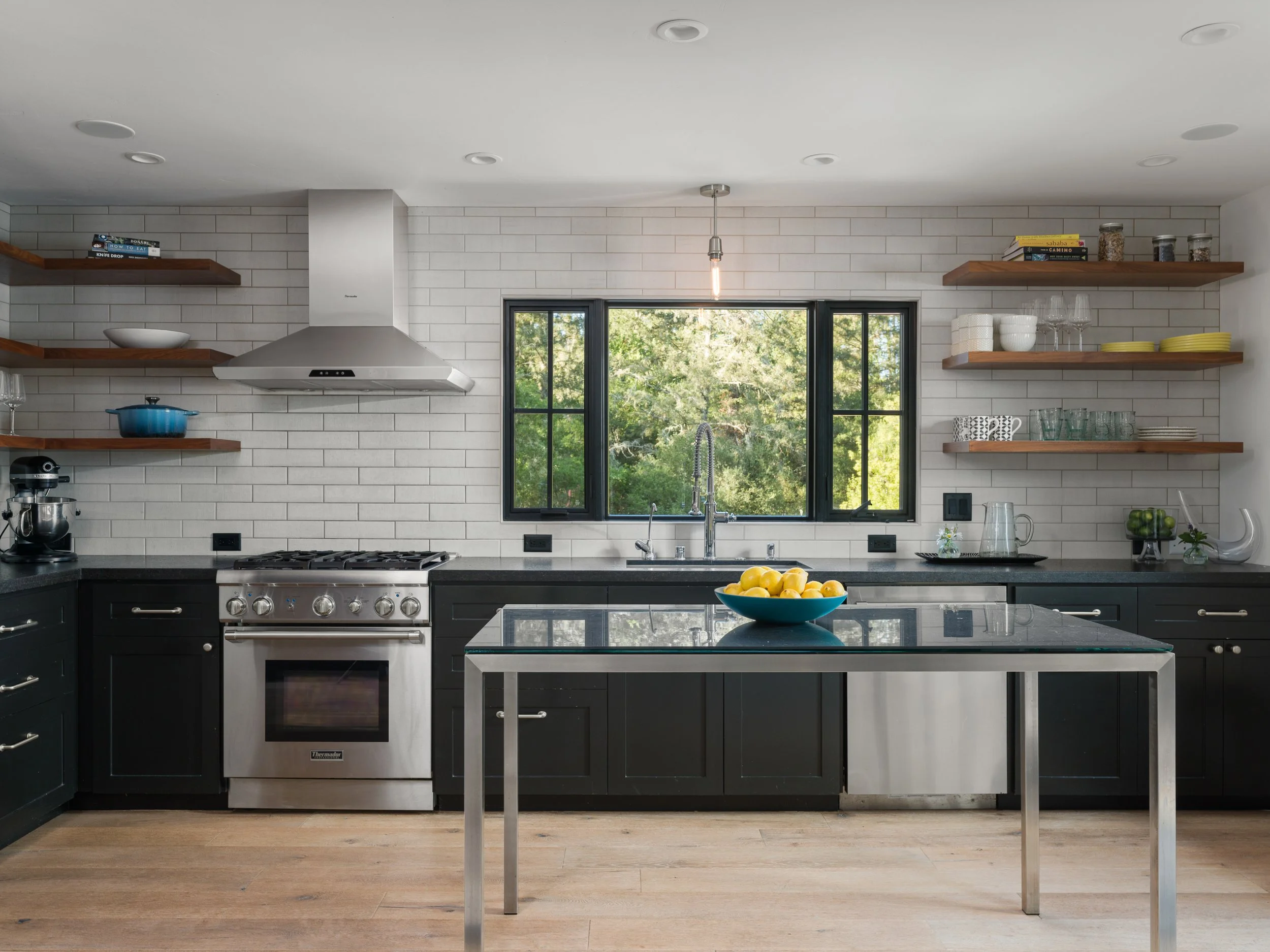 Modern kitchen with white tile walls, black cabinets, stainless steel stove, and open wooden shelves holding books, bowls, glasses, and jars. Large window shows green trees outside.