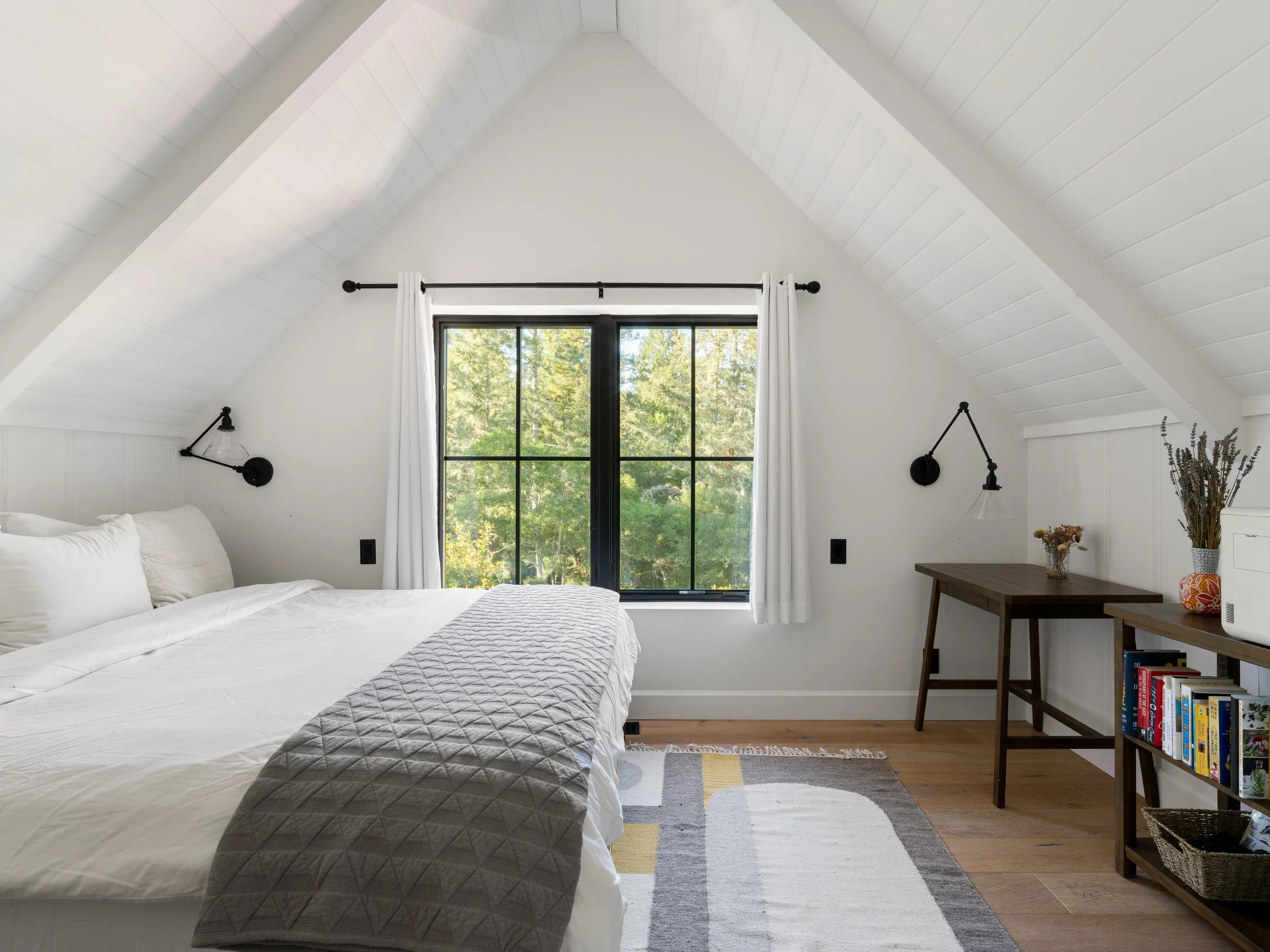 A cozy bedroom with a white bed, gray quilted blanket, large window with white curtains, wall-mounted black lights, a wooden desk with a vase of dried flowers, a small bookshelf with books, and a colorful geometric area rug.