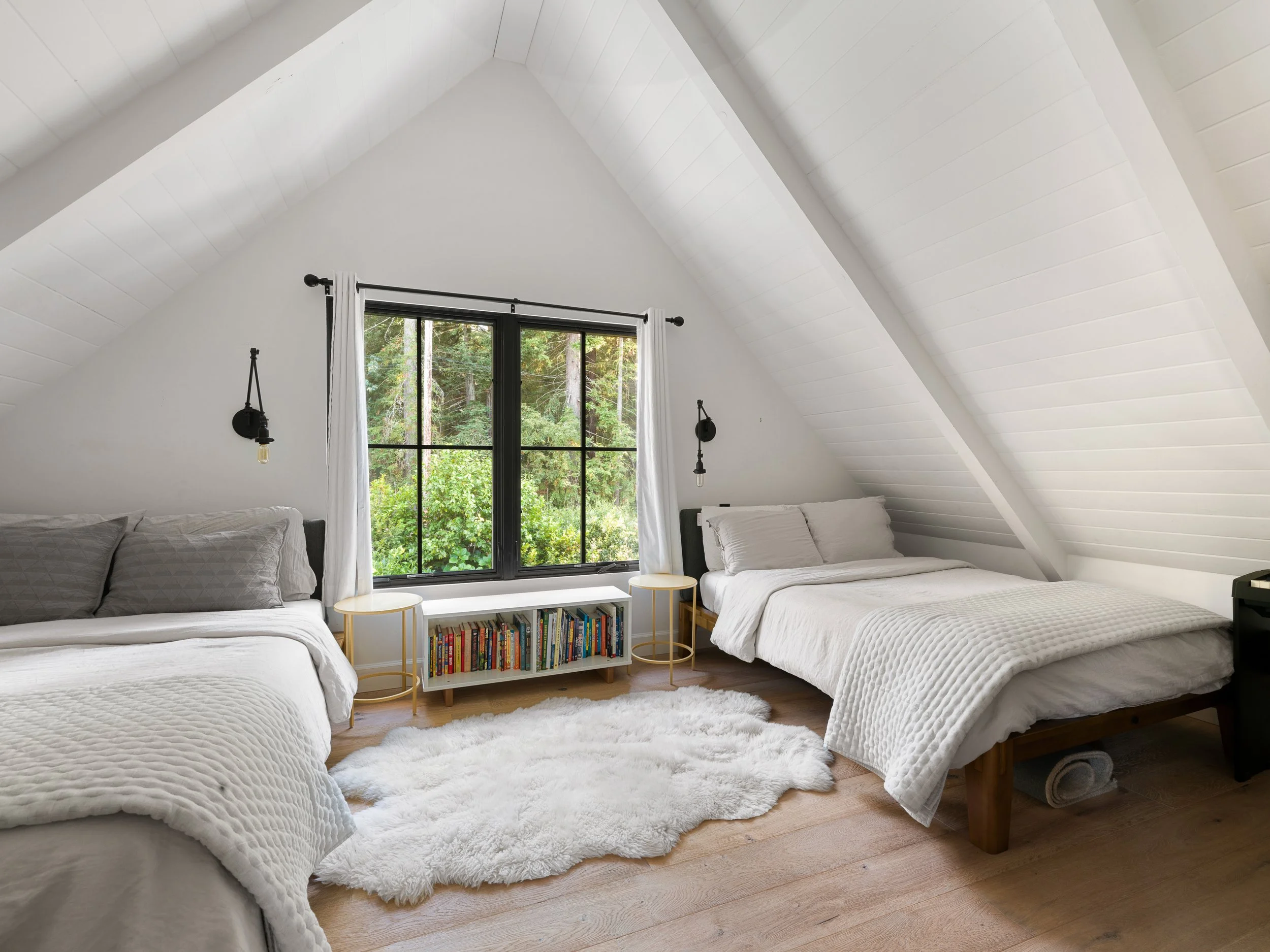 A cozy bedroom with white walls, a vaulted ceiling, and a large window showing green trees outside. There are two beds with white bedding, a white shaggy rug on the wooden floor, and a small white bookshelf filled with books between the beds. Black w