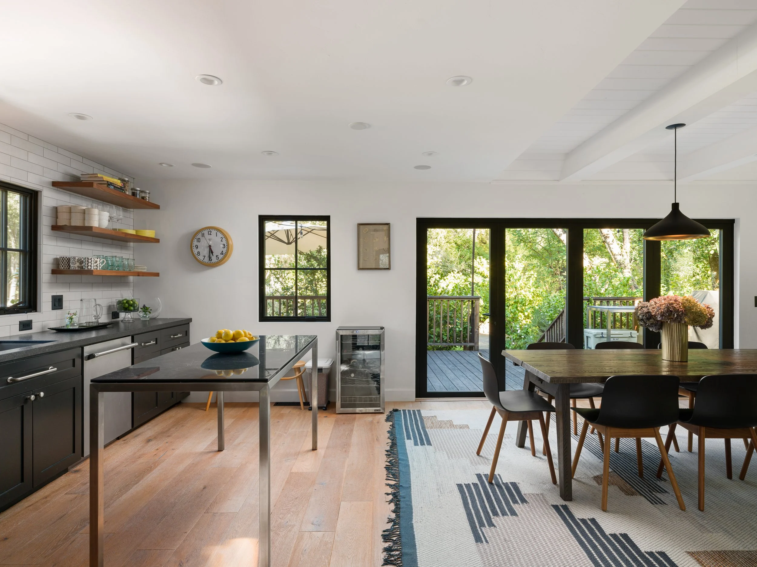 Open-concept kitchen and dining area with black cabinetry, gray range, wooden open shelves, a wall clock, large windows, a dining table with black chairs, and a sliding glass door leading to an outdoor deck surrounded by trees.