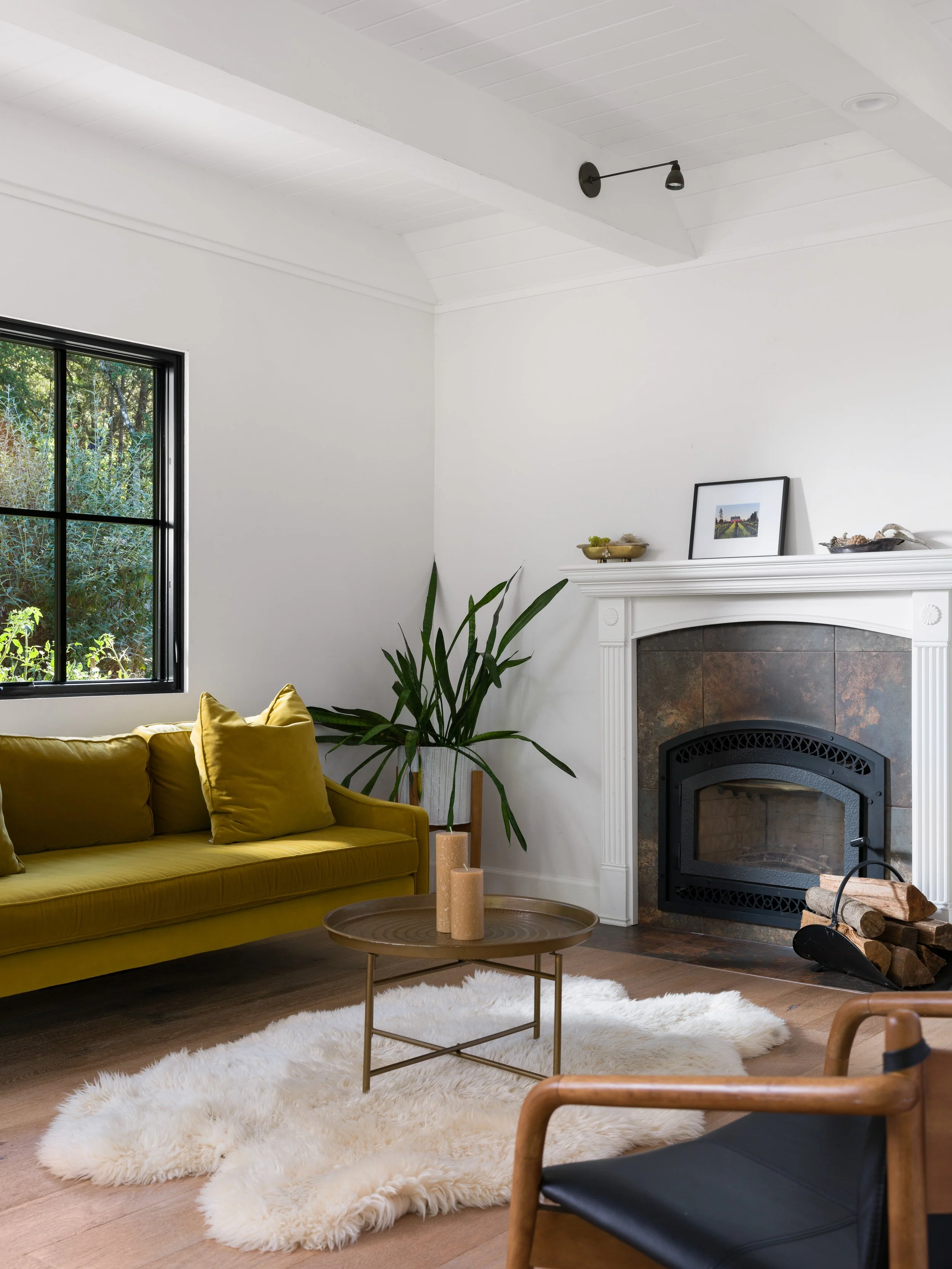 Living room with a yellow sofa, green plant, fireplace with framed photo and decorative items, window with greenery outside, white shaggy rug, wooden floor, and wooden furniture.