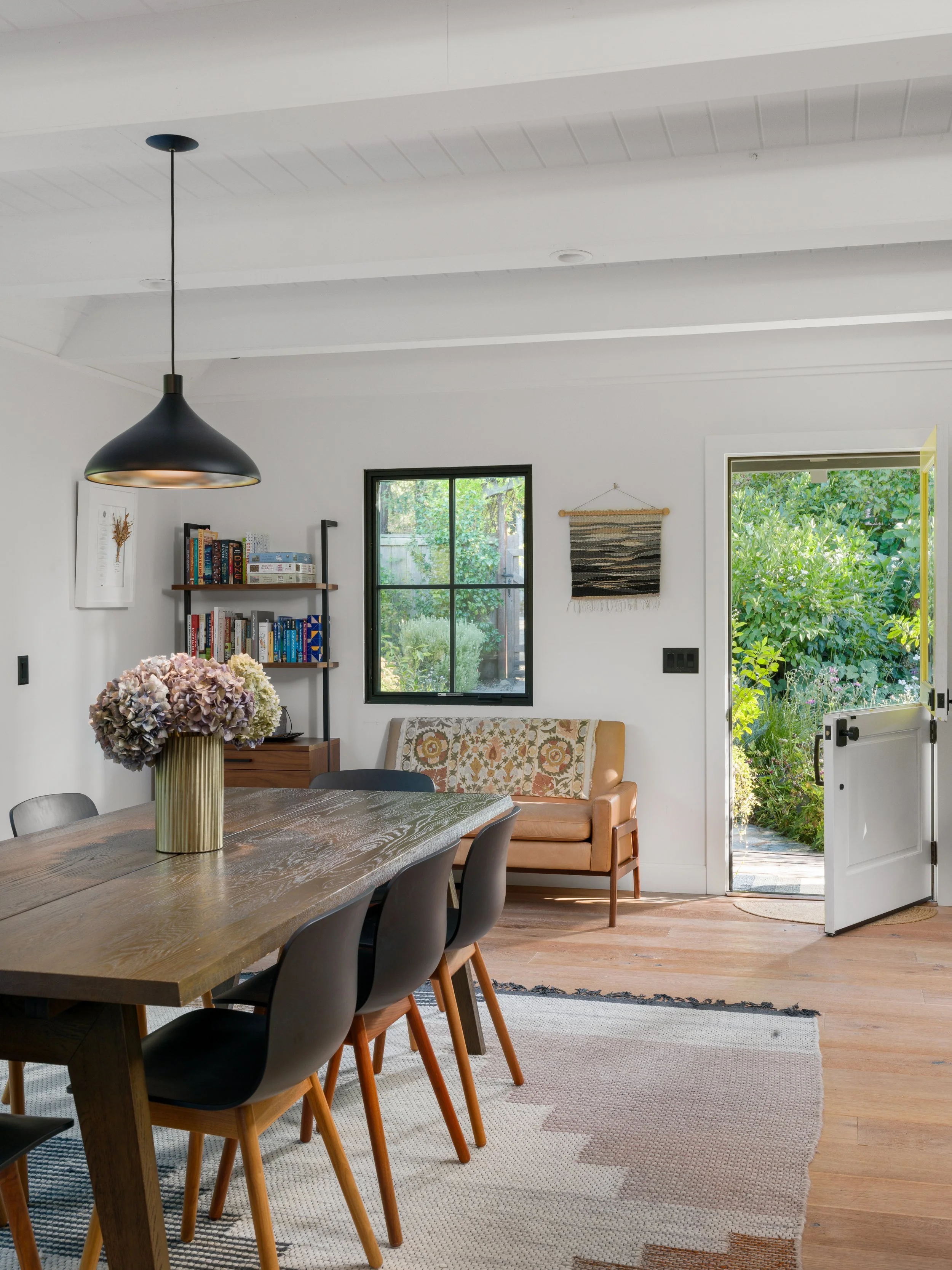 A cozy dining room with a wooden table, black chairs, a bouquet of flowers, and a view of greenery outside through an open door and window, with a bookshelf on the wall and a wall hanging.