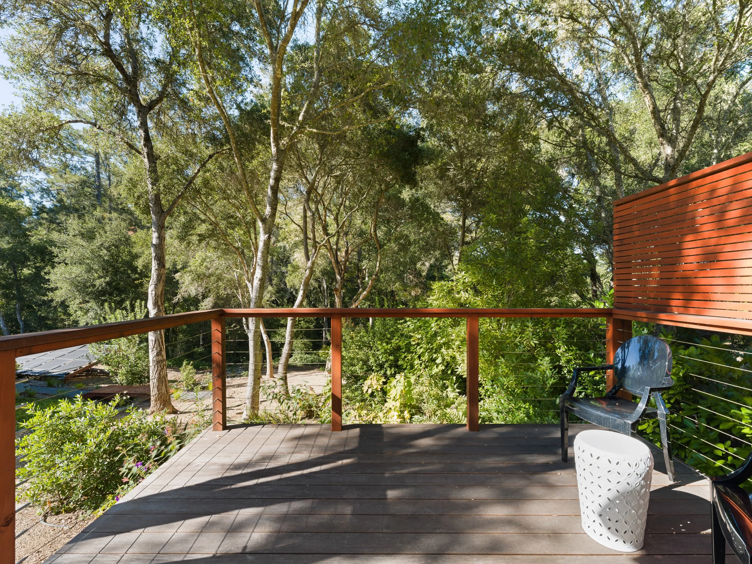 A view of an outdoor wooden deck with patio furniture, including a glass chair, a white decorative planter, and lush green trees in the background, with sunlight filtering through the leaves.