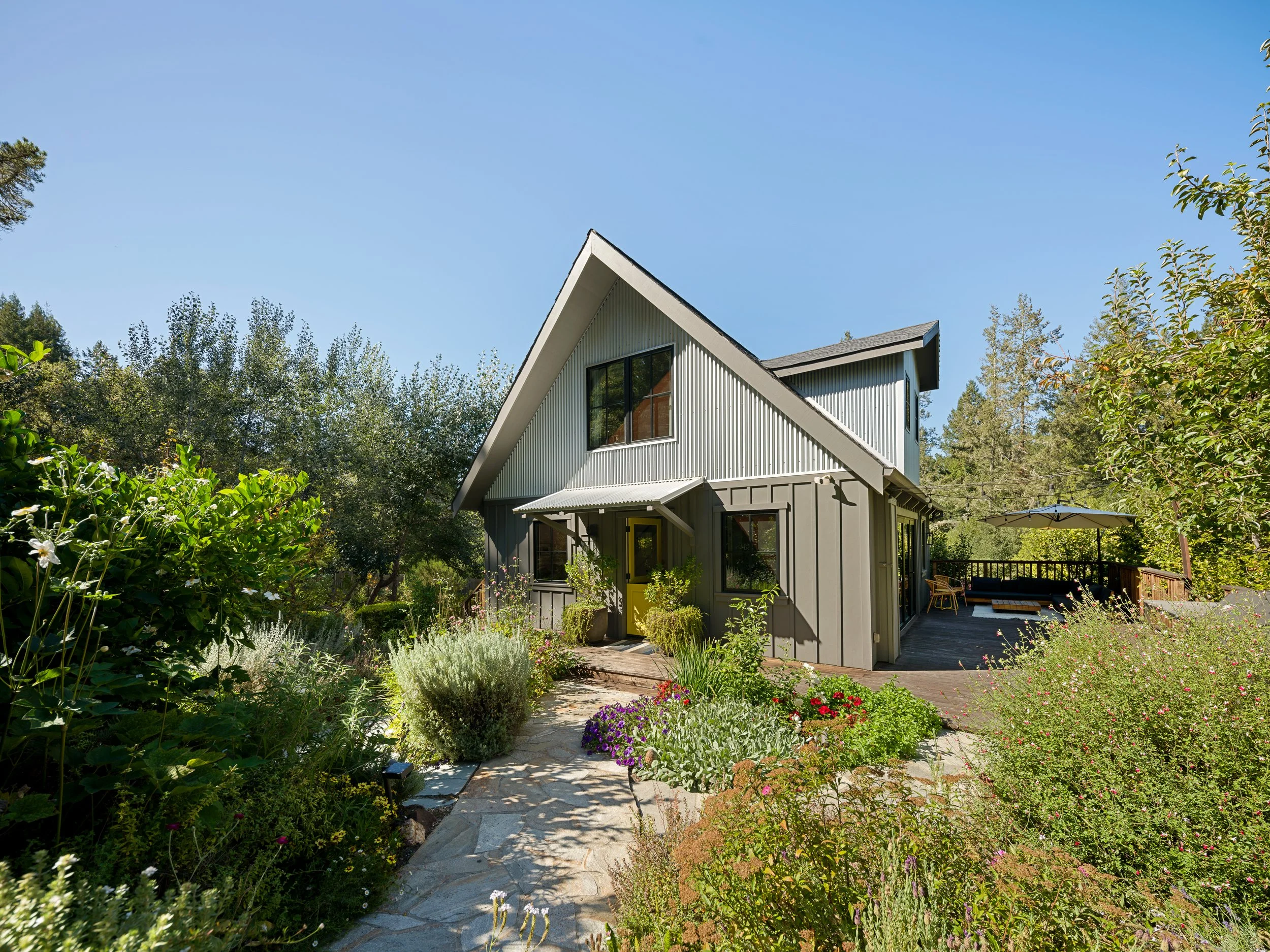 A house with a steep gabled roof and large windows, surrounded by lush garden with various flowers and greenery, a stone pathway, and a patio with an umbrella.
