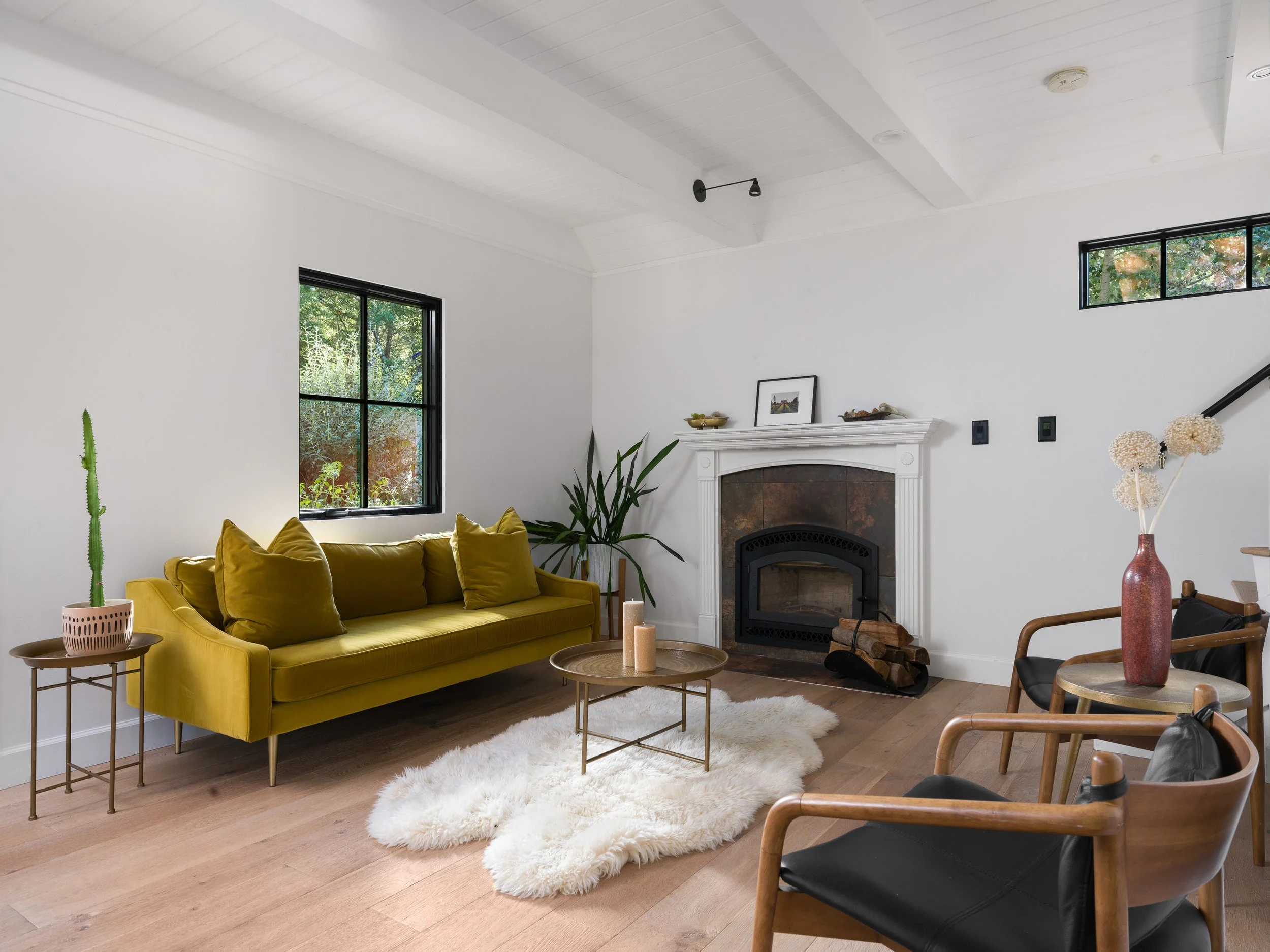 A living room with a mustard yellow sofa, green plants, a white fluffy rug, a fireplace, wooden floors, and various decorative items in the room.