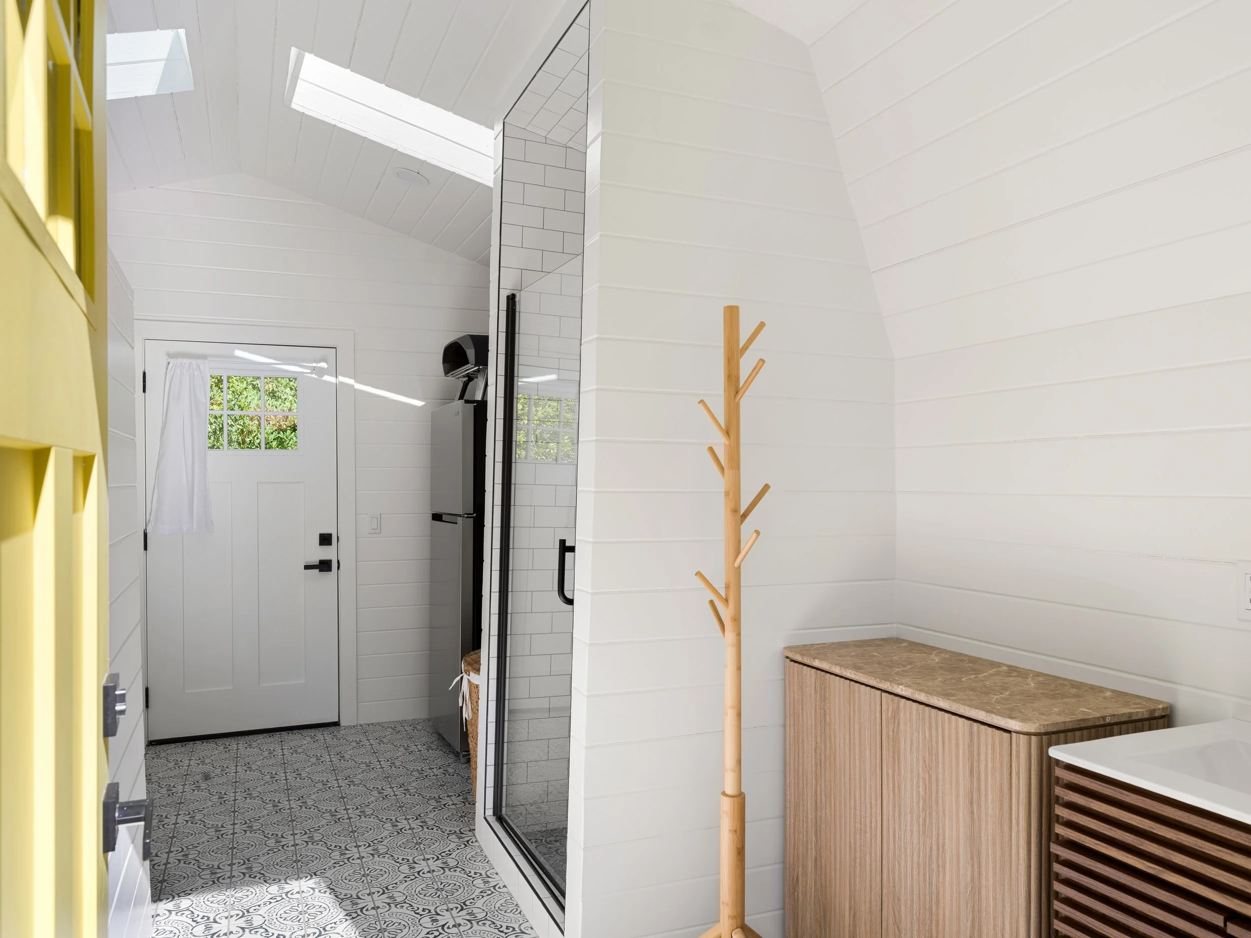 Interior view of a bright, modern entryway with a white paneled ceiling, a skylight, patterned tile floor, white wall paneling, a wooden coat rack, and a brown cabinet.
