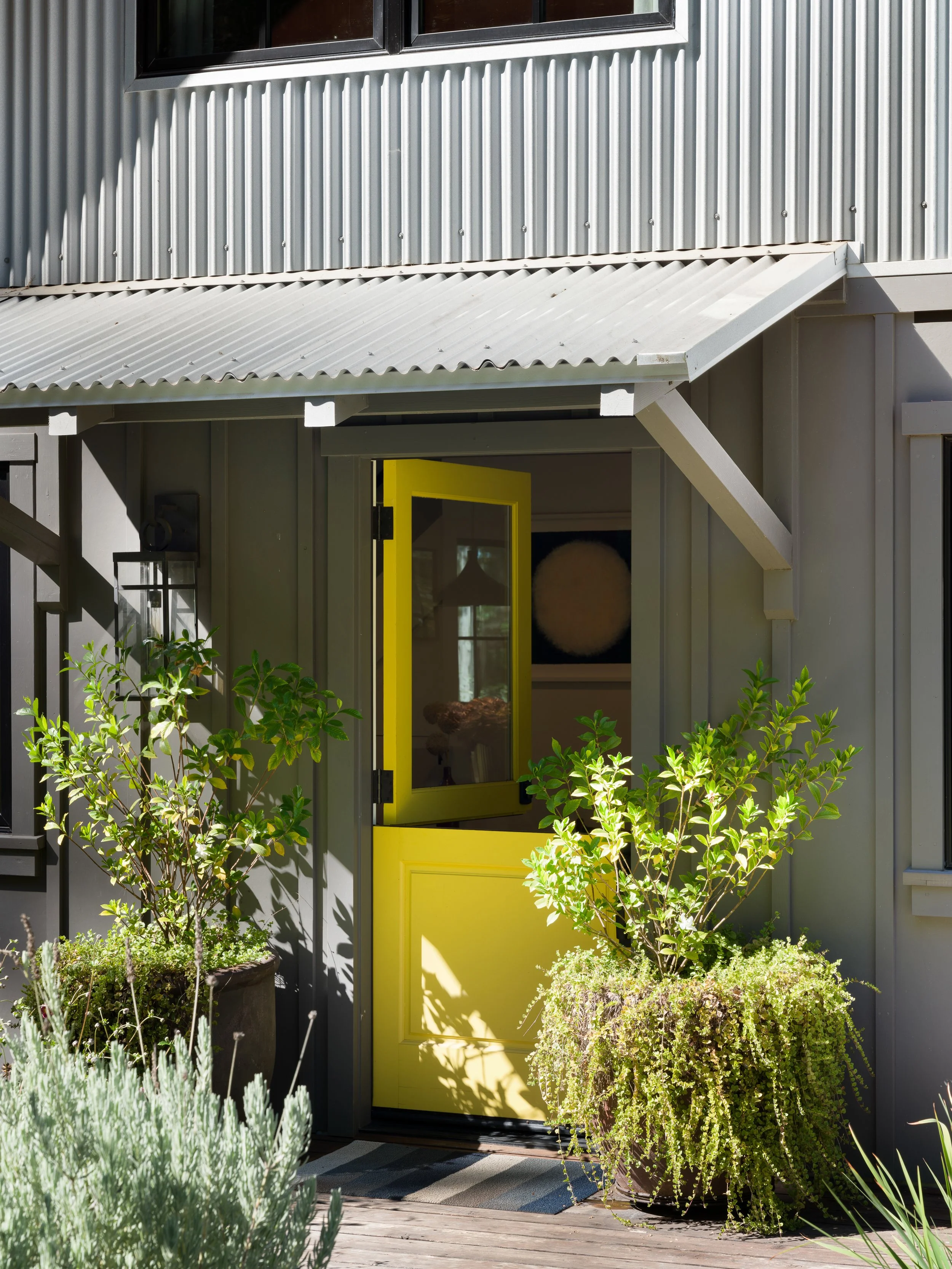 A yellow door with a glass panel opens onto a small porch with potted plants and greenery in the foreground. The building has gray vertical siding and a corrugated metal awning above the door, with a window above.