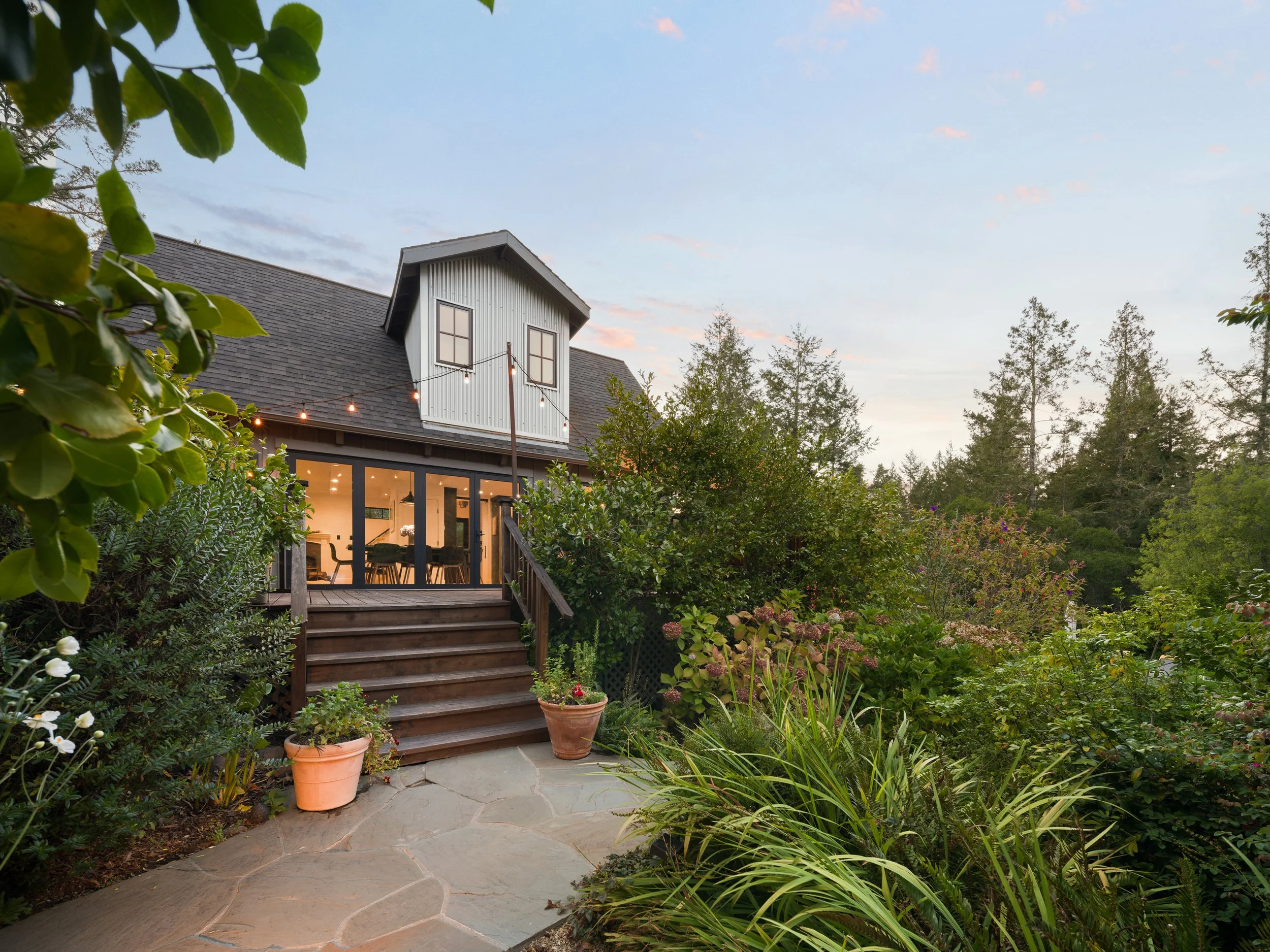 A backyard garden with a stone pathway, potted plants, lush greenery, and a house with a deck and string lights in the background, with trees and a clear sky.