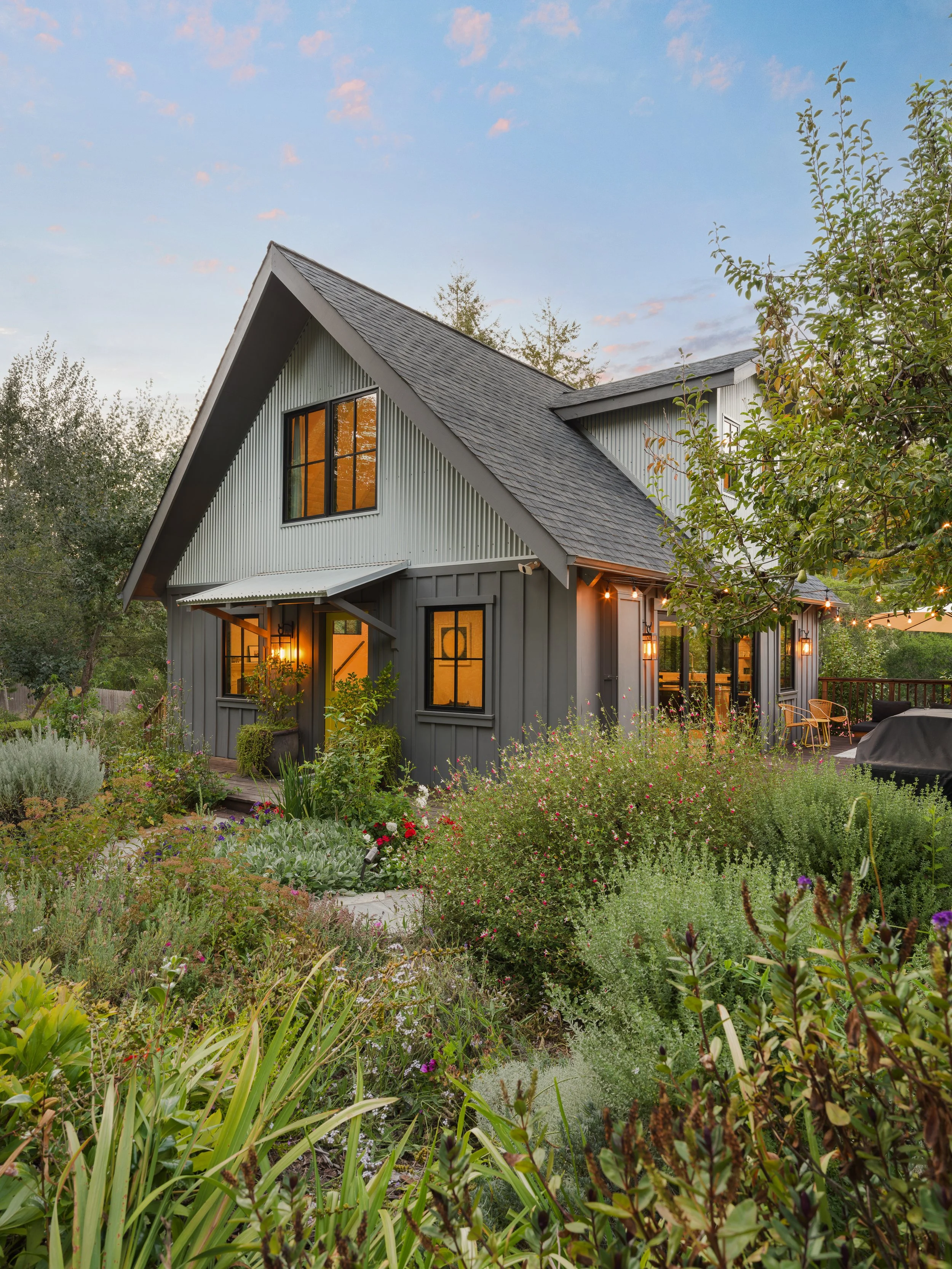 A modern house with large windows, a steep gabled roof, and an inviting outdoor porch surrounded by lush garden plants