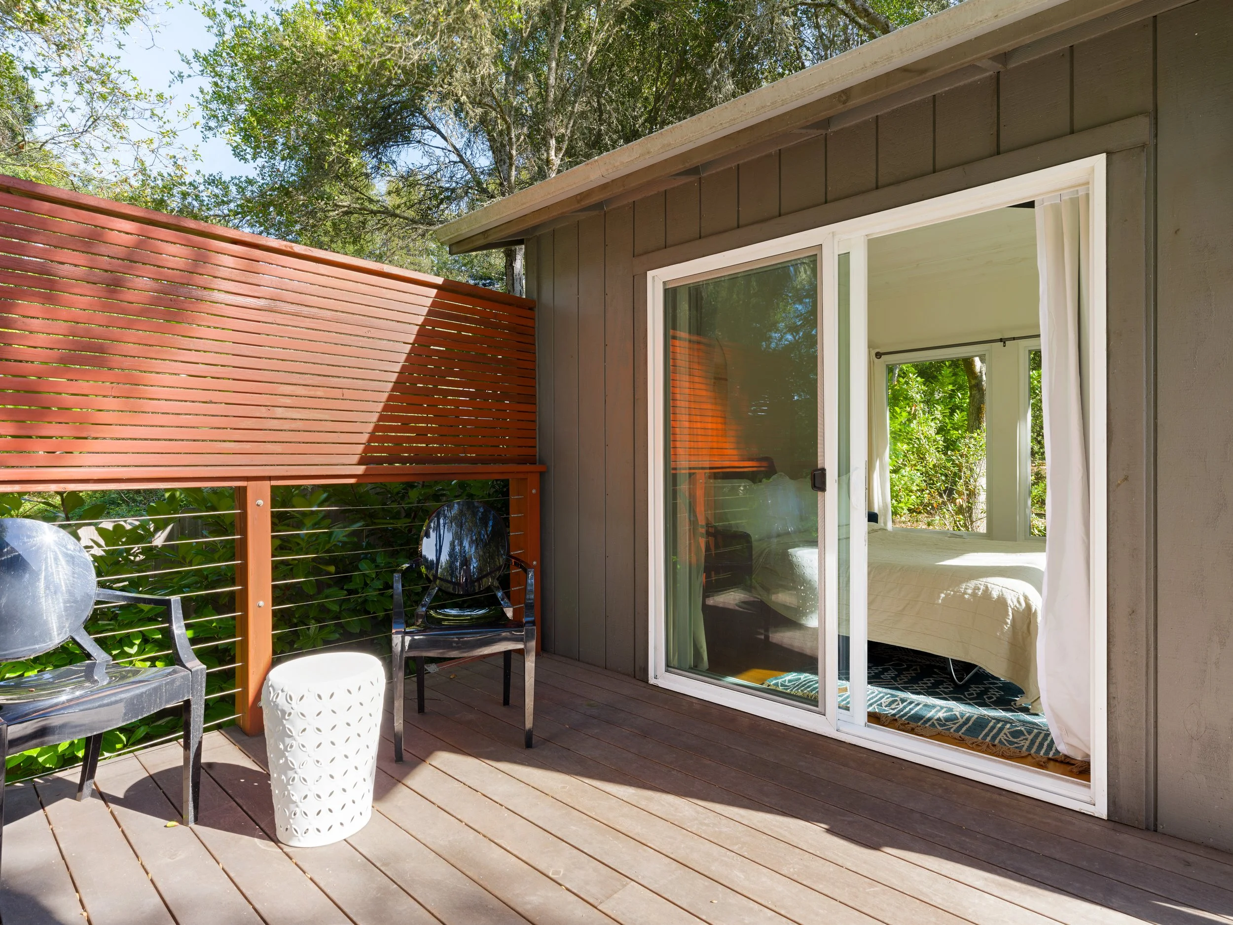 Patio with wooden deck, black chairs, white side table, and sliding glass door leading to indoor bedroom with bed and blue rug, surrounded by green trees and a reddish-brown wooden privacy screen.