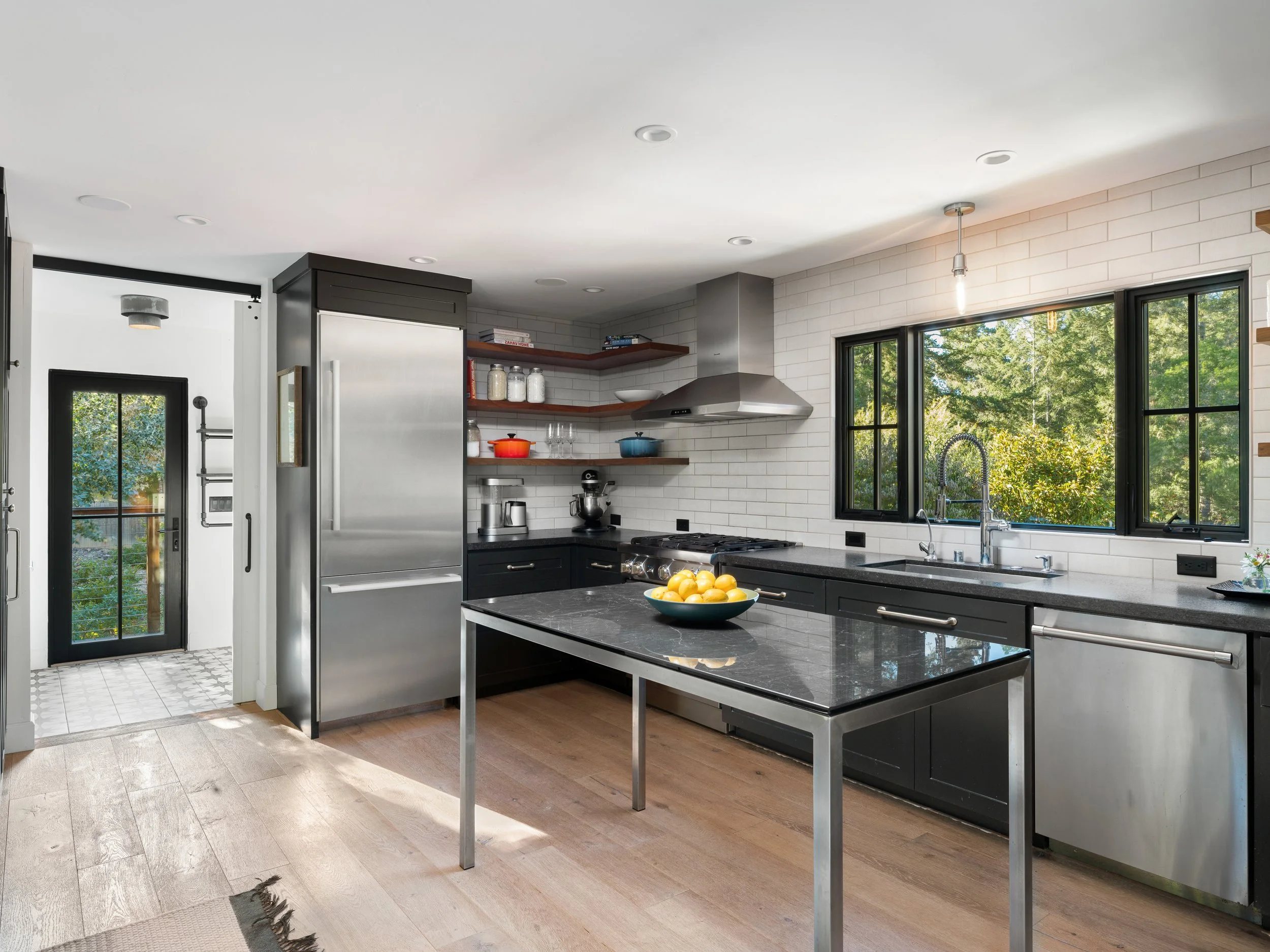 Modern kitchen with black cabinets, white subway tile backsplash, open wooden shelves, stainless steel appliances, large windows showing green trees outside, black-framed door, and a kitchen island with a bowl of yellow lemons.