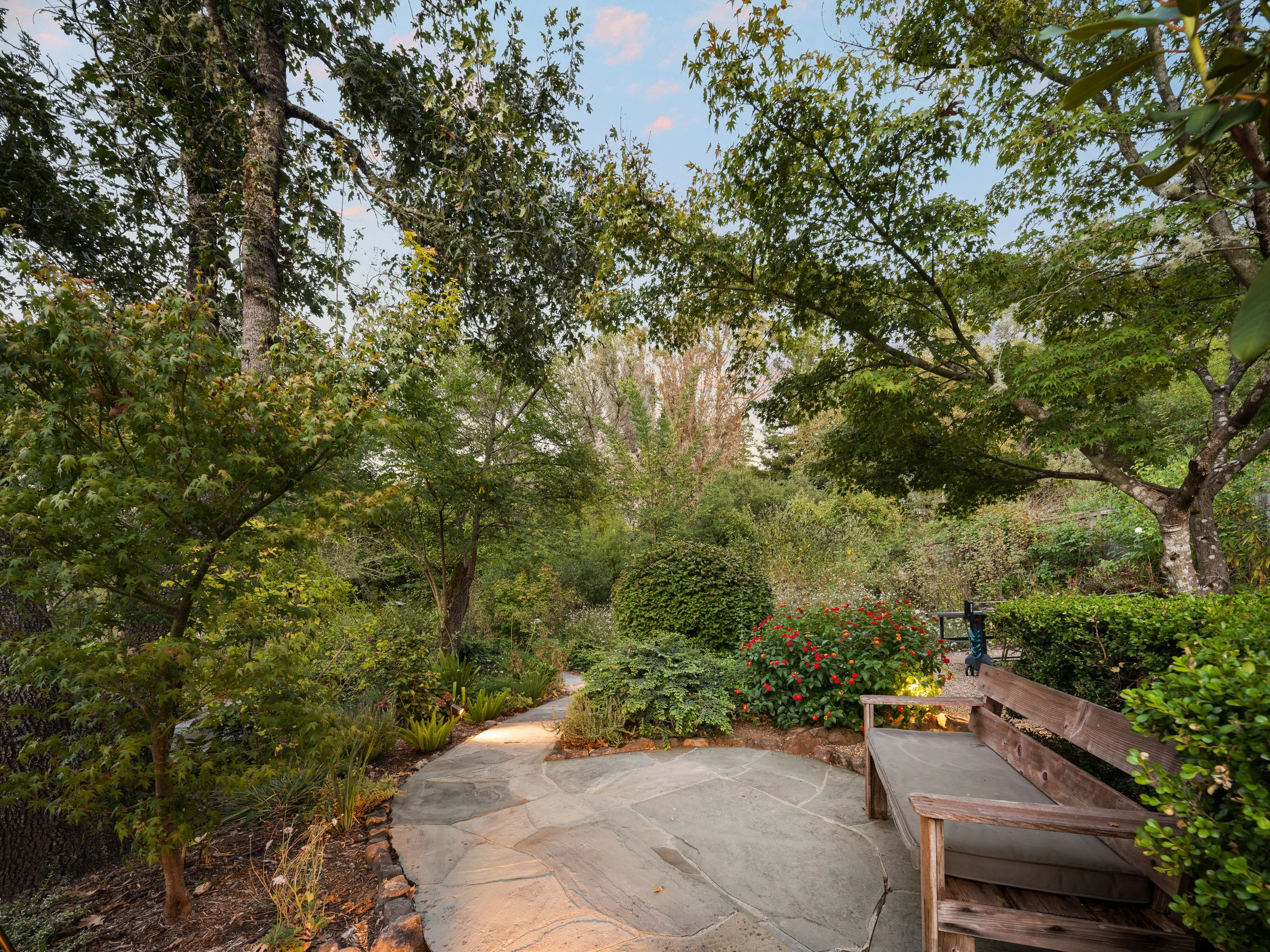A backyard garden with a stone pathway, surrounded by lush green trees and bushes; a wooden bench with gray cushion on the right side of the image.