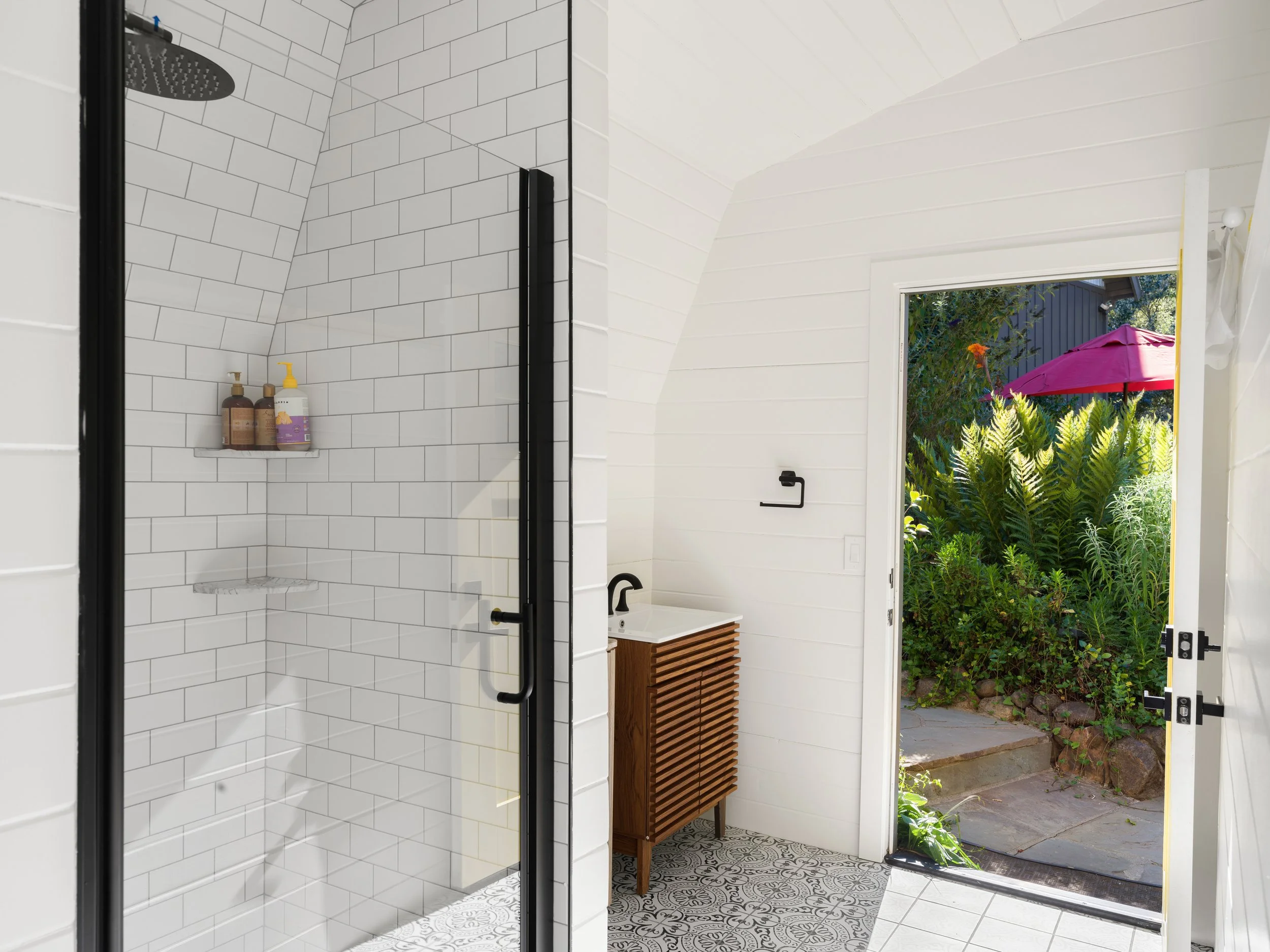 View from inside a shower with white subway tiles, overlooking a door leading outside to a garden with plants and a pink umbrella.