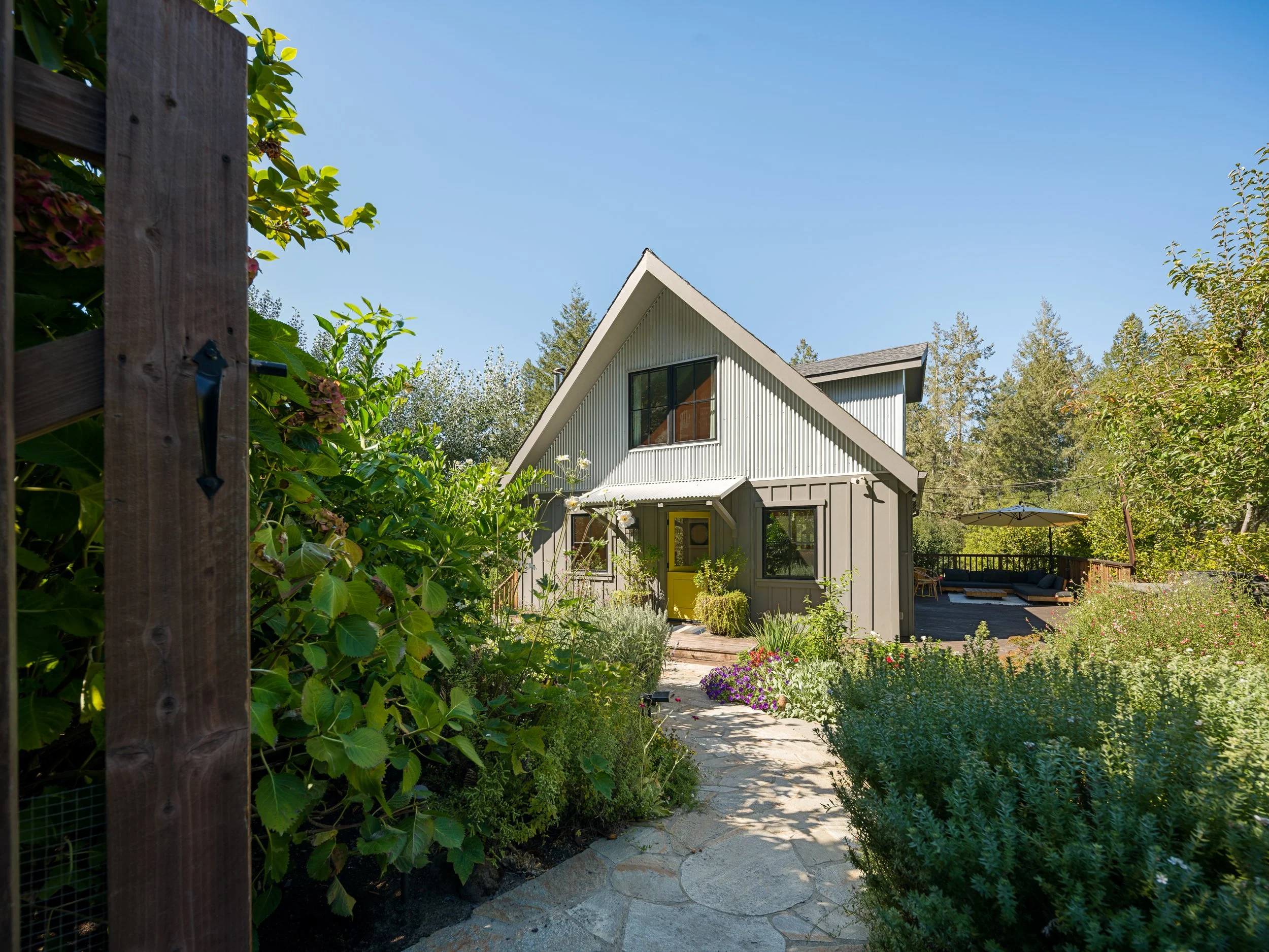 A modern house with a triangular roof and large windows, surrounded by a lush garden with various flowers and plants, with a clear blue sky overhead.
