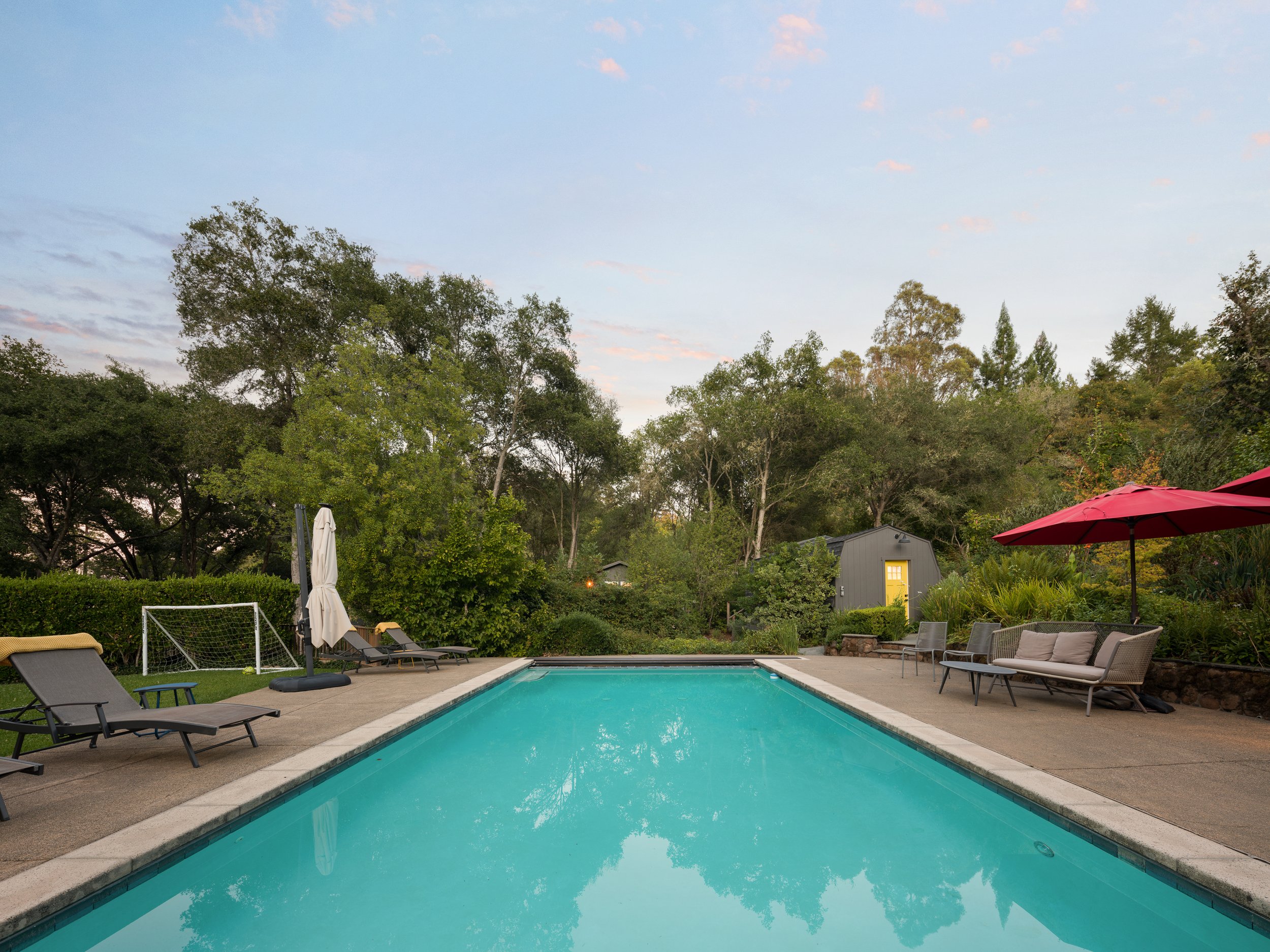 A backyard scene with a swimming pool, lounge chairs, umbrellas, lush trees, a small gray building, and a partly cloudy sky.