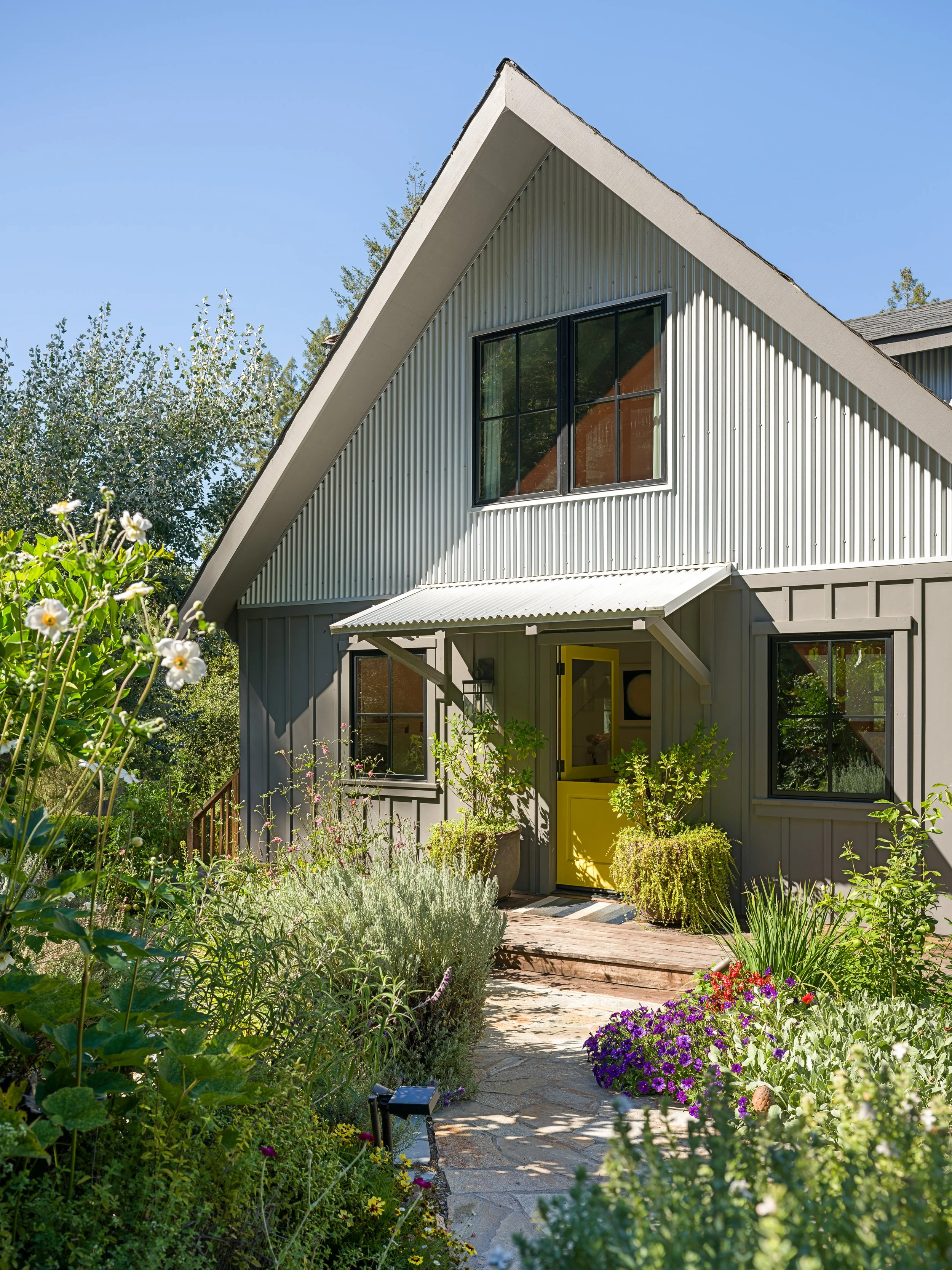 A modern house with a steeply sloped roof and a large window, surrounded by a lush garden with various flowers and greenery on a bright, sunny day.