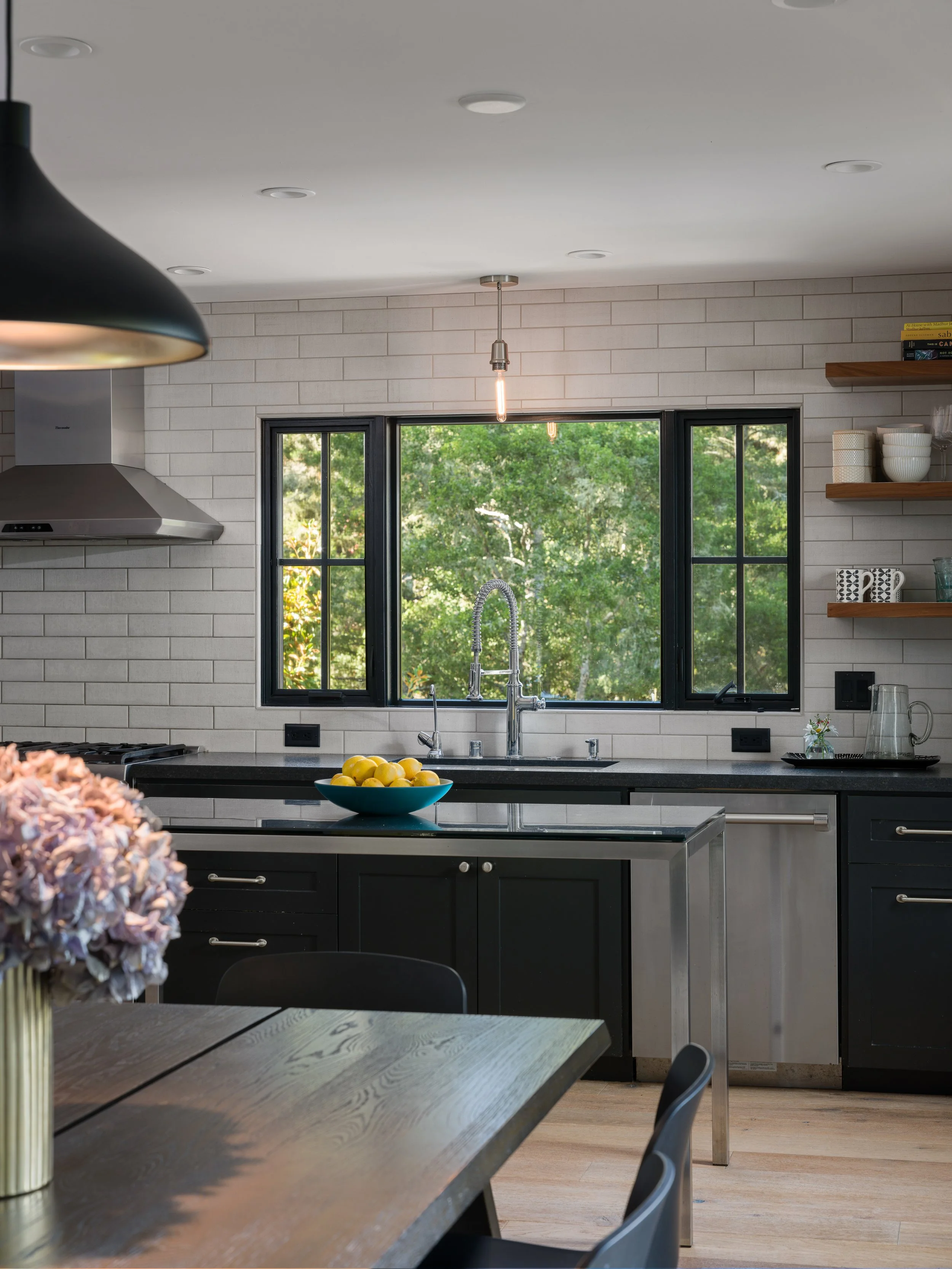 Modern kitchen with black cabinets, a dark countertop, stainless steel appliances, a window showing greenery outside, and a dining table with a vase of flowers.