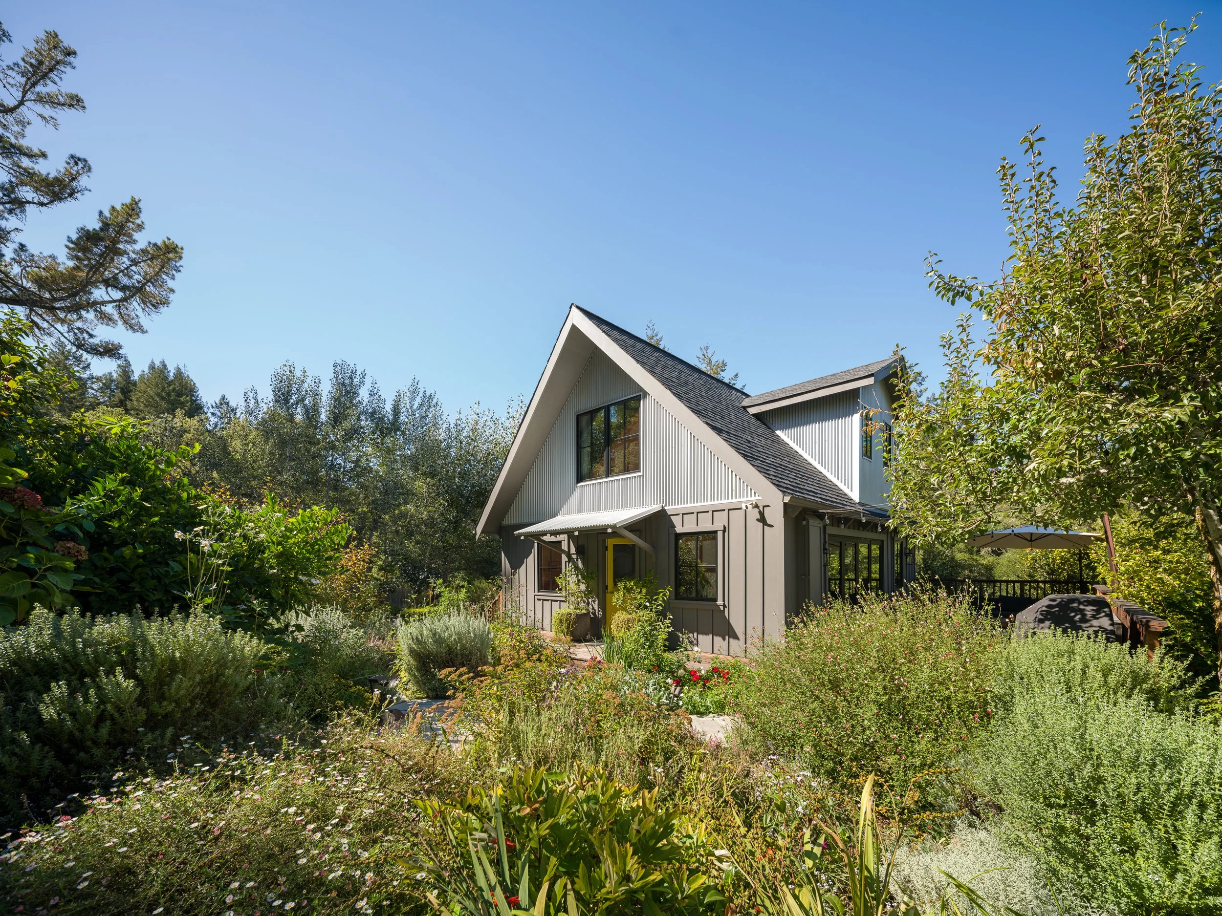 A white house with a steep roof is surrounded by a lush garden with various plants, shrubs, and trees under a clear blue sky.