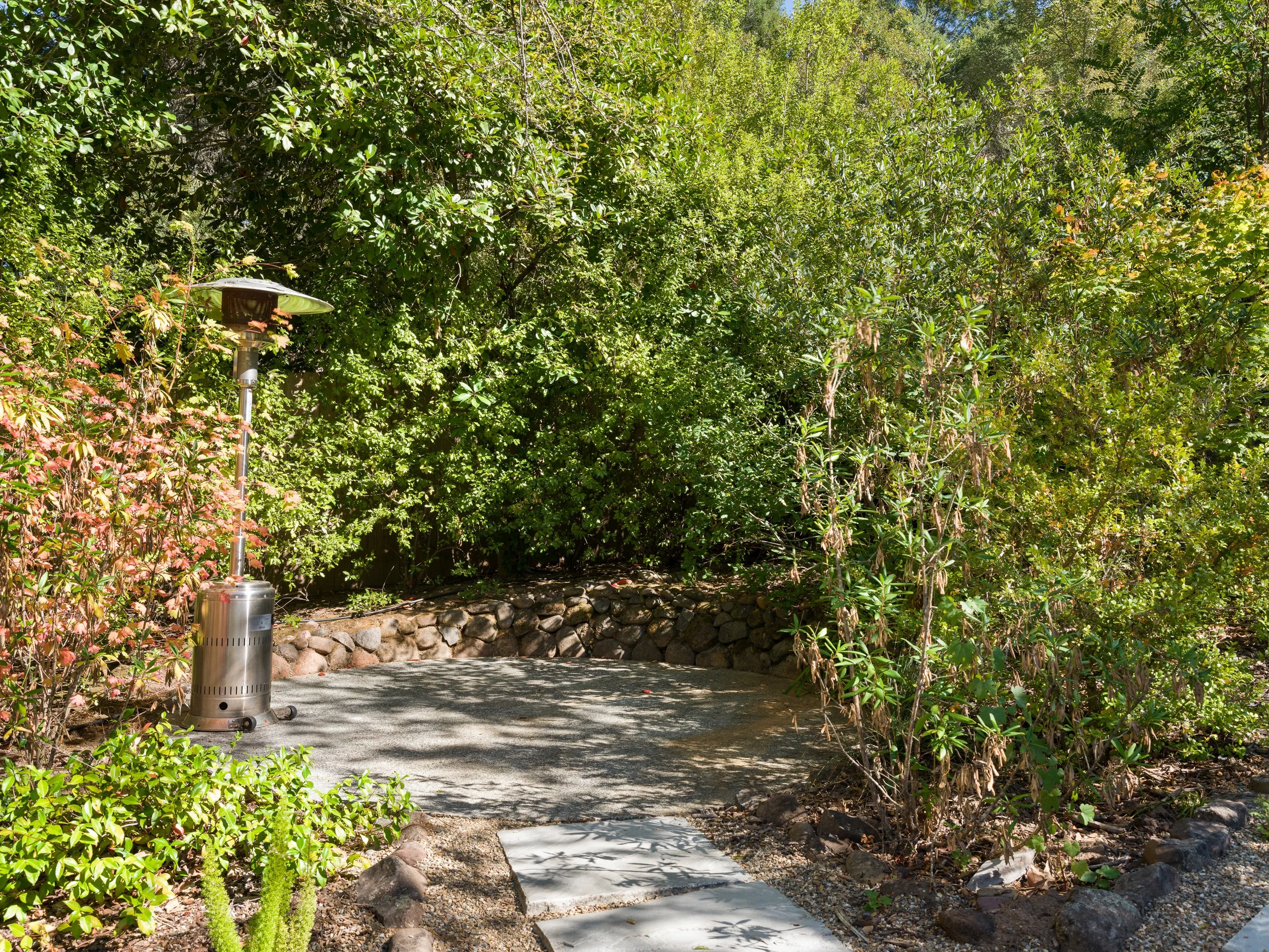 A small concrete sidewalk curves through a lush, green garden with dense foliage on both sides. There is a portable outdoor heater on the left side of the path, and sunlight filters through the leaves, creating dappled shadows on the ground.
