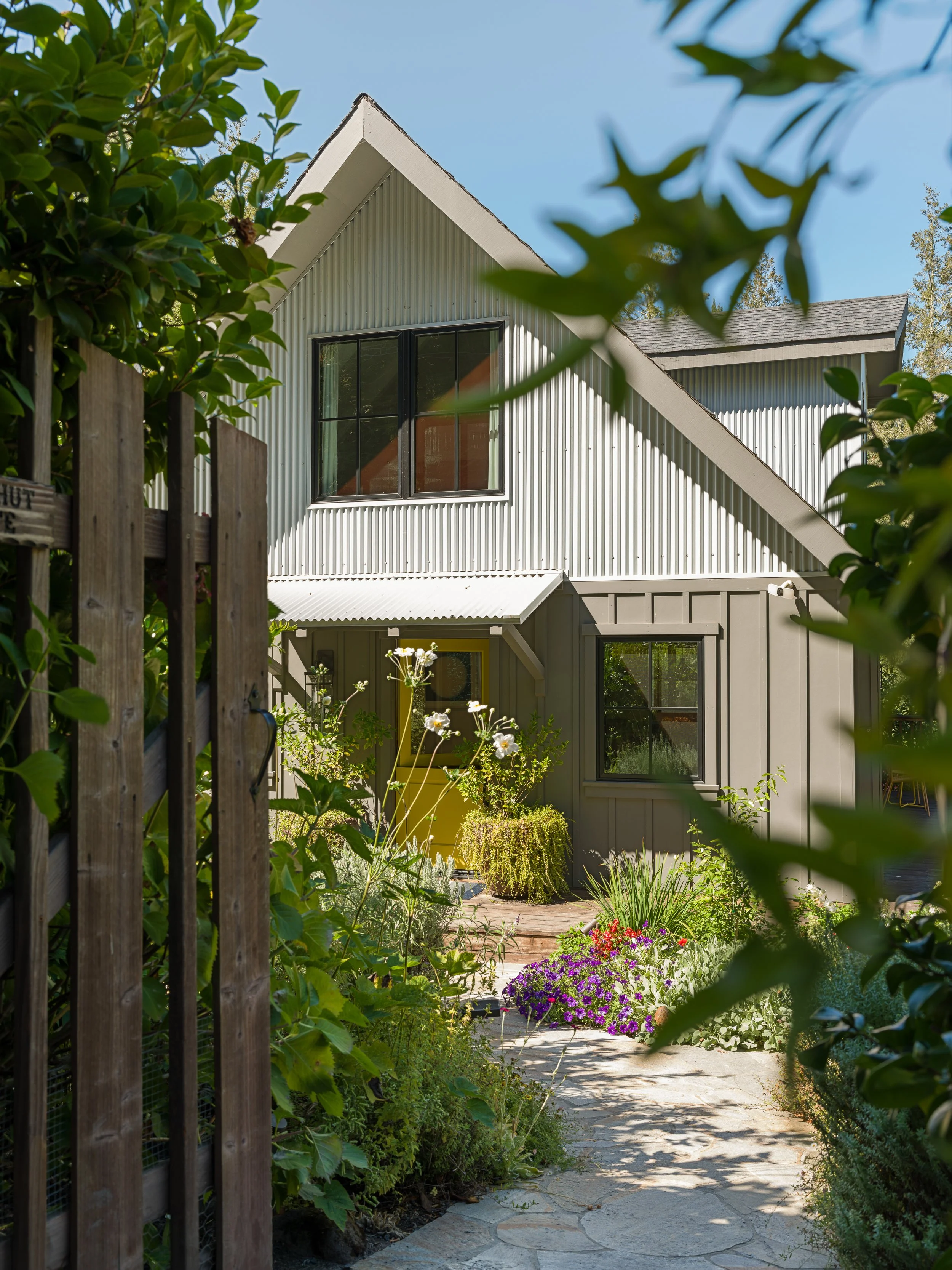 A backyard garden entrance with a stone pathway, surrounded by colorful flowers and lush green plants, leading to a modern house with metal siding and large windows.