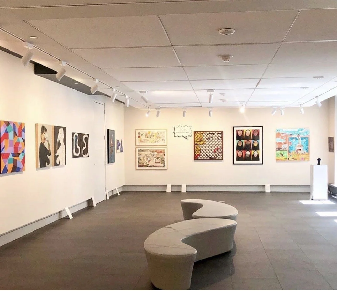 Art gallery with colorful paintings on white walls, modern curved white bench in the center, artwork illuminated by track lighting, vent on ceiling, light-colored flooring, and a small pedestal with a sculpture.