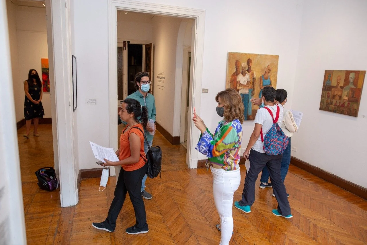 People viewing paintings at an art gallery, all wearing masks, with some engaging with the artwork and a few taking photos.