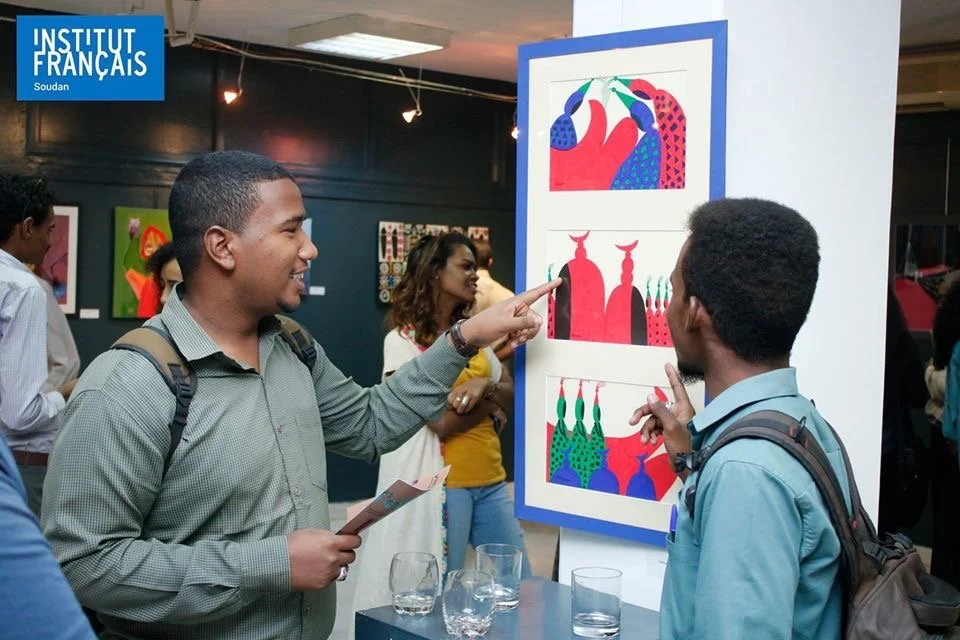 Two men are viewing colorful modern art prints at an art exhibit, engaging in conversation, with other attendees and artworks in the background.