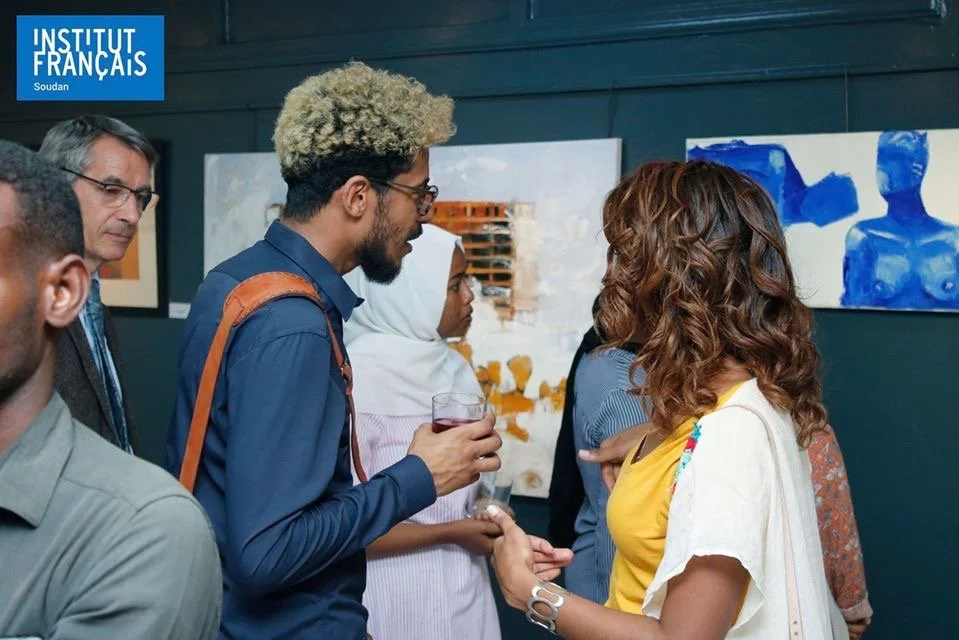 A diverse group of people at an art gallery, including a woman shaking hands with a man holding a drink, while others look at paintings on the wall.