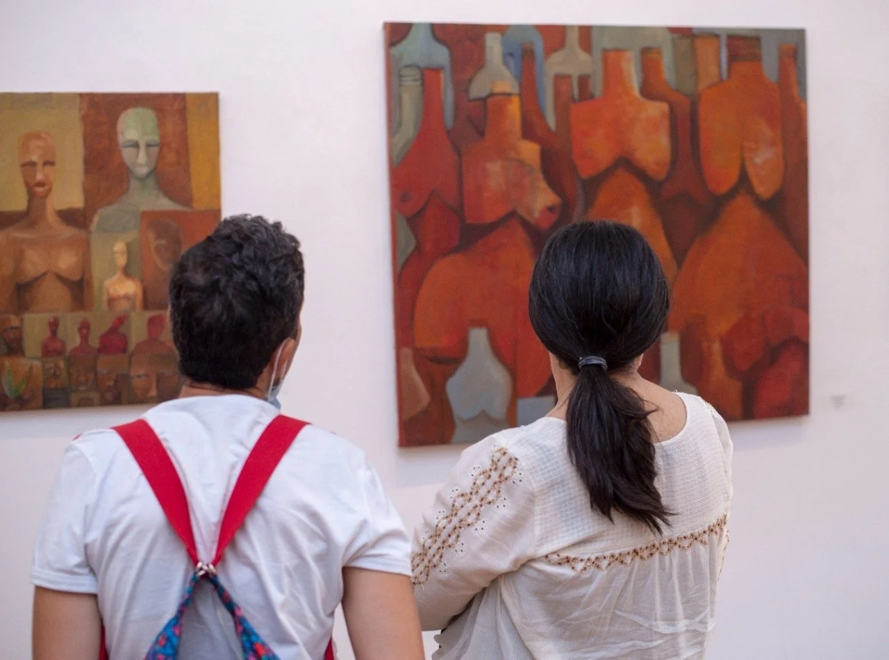 Two people standing in front of colorful abstract paintings in an art gallery. One person has dark hair tied back, and the other has short dark hair wearing a mask and red suspenders.