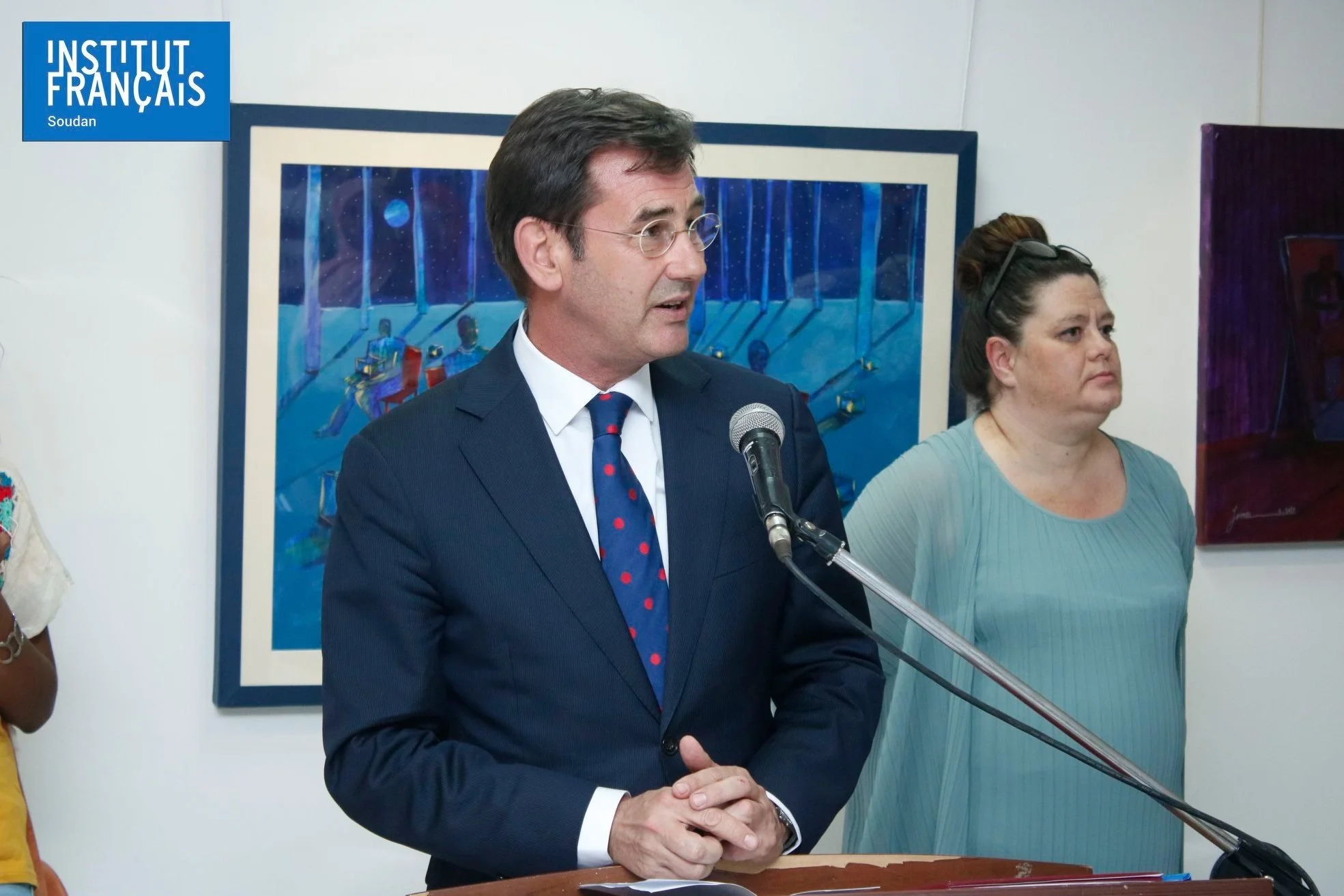 Man in a dark suit and glasses speaking at a microphone, standing in front of a colorful abstract painting, with a woman in a light blue top standing behind him.