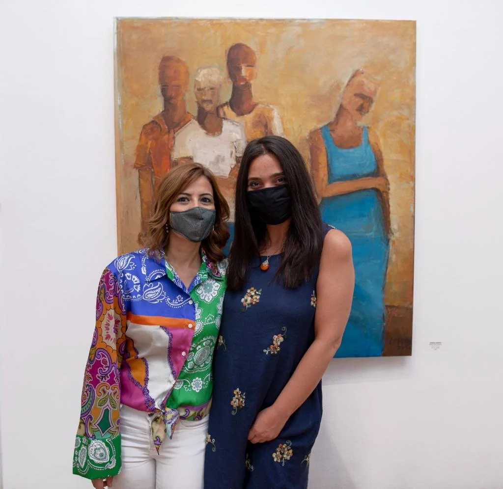 Two women standing in front of an artwork in a gallery, both wearing masks and colorful dresses.