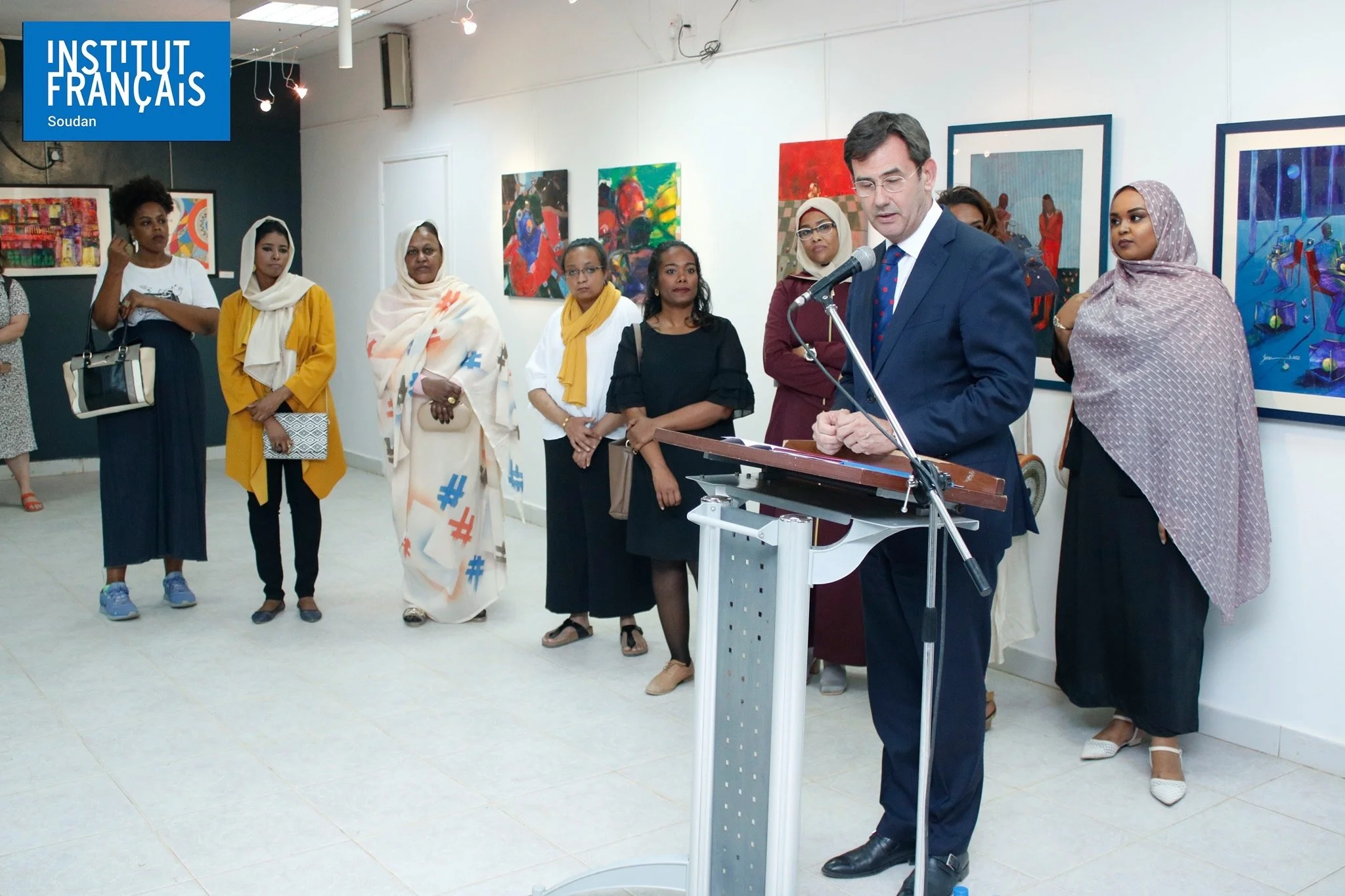 A group of diverse women listening to a man in a suit giving a speech at an art exhibition, with colorful paintings on the wall behind them.