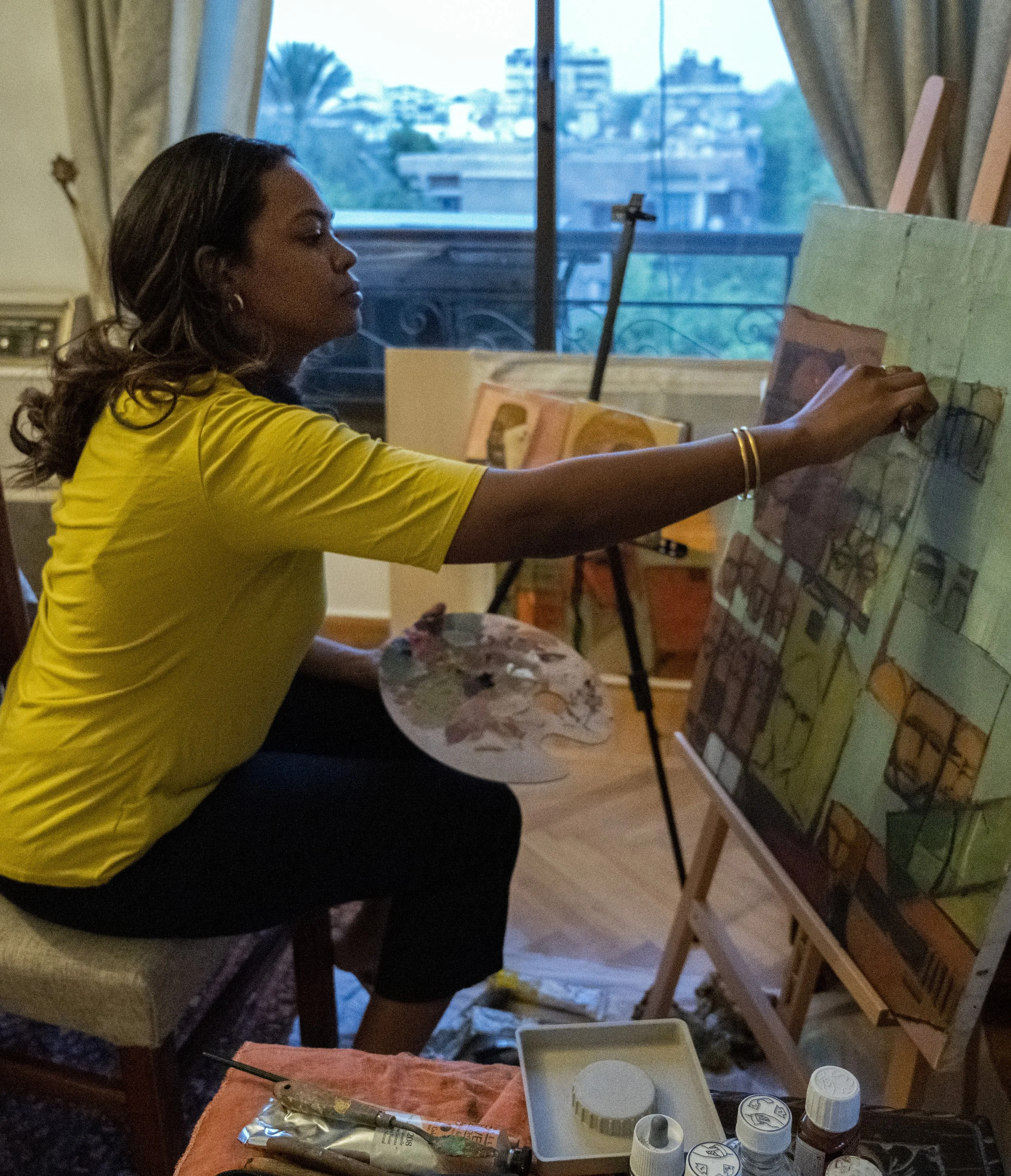 A woman in a yellow shirt is sitting at an easel, painting a landscape scene with a cityscape. She is holding a paintbrush, and various painting supplies, including tubes of paint and containers, are on the table in front of her. A large window with curtains reveals a view of buildings and trees outside.