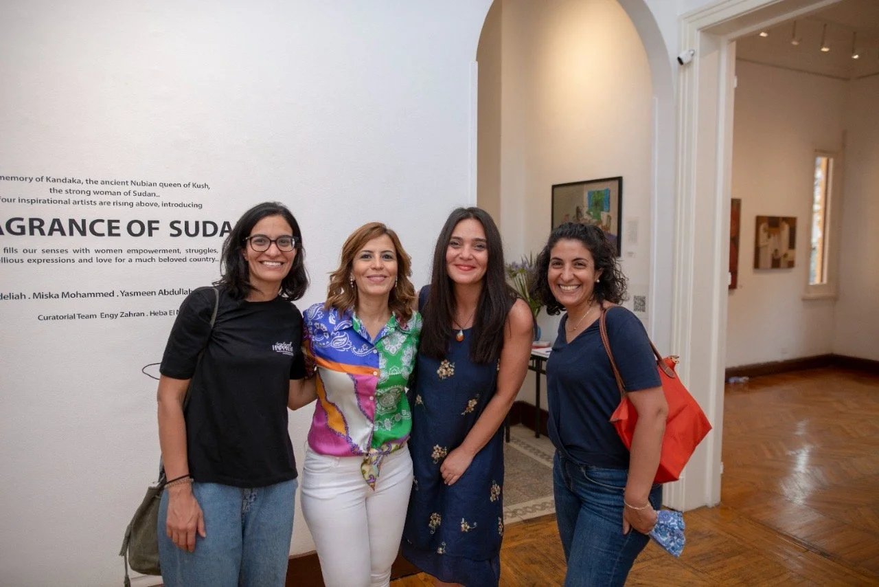 Four women standing together in an art gallery, smiling at the camera, with artwork on the walls behind them.