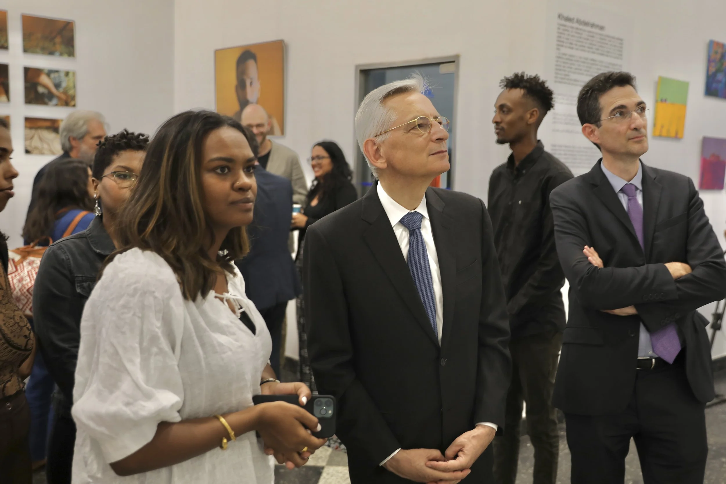 A diverse group of people attending an art exhibition, observing artworks and listening attentively, with a woman in white holding a phone in the foreground.