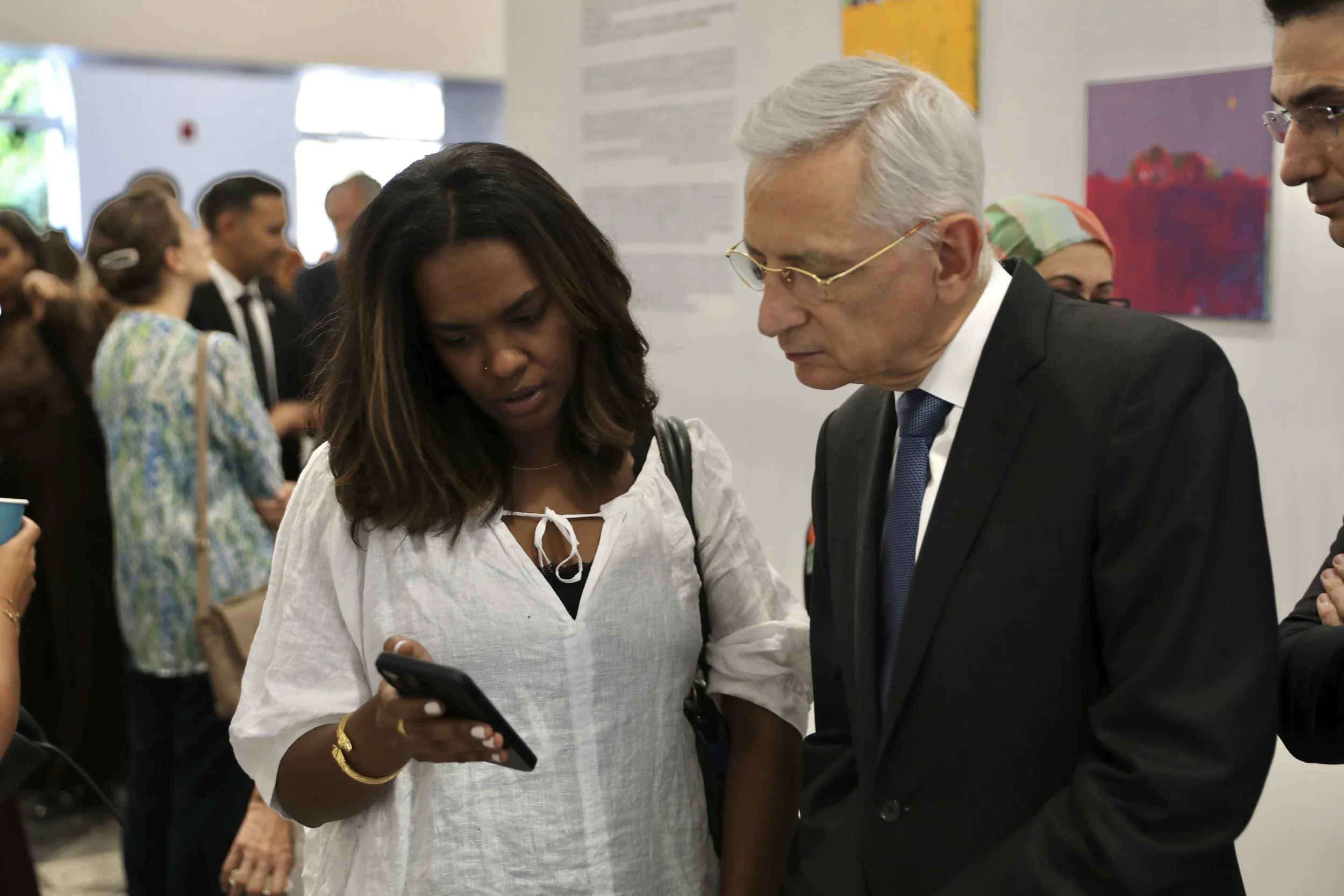 A woman and a man in formal attire are looking at a smartphone together at an art gallery, with several other people in the background.