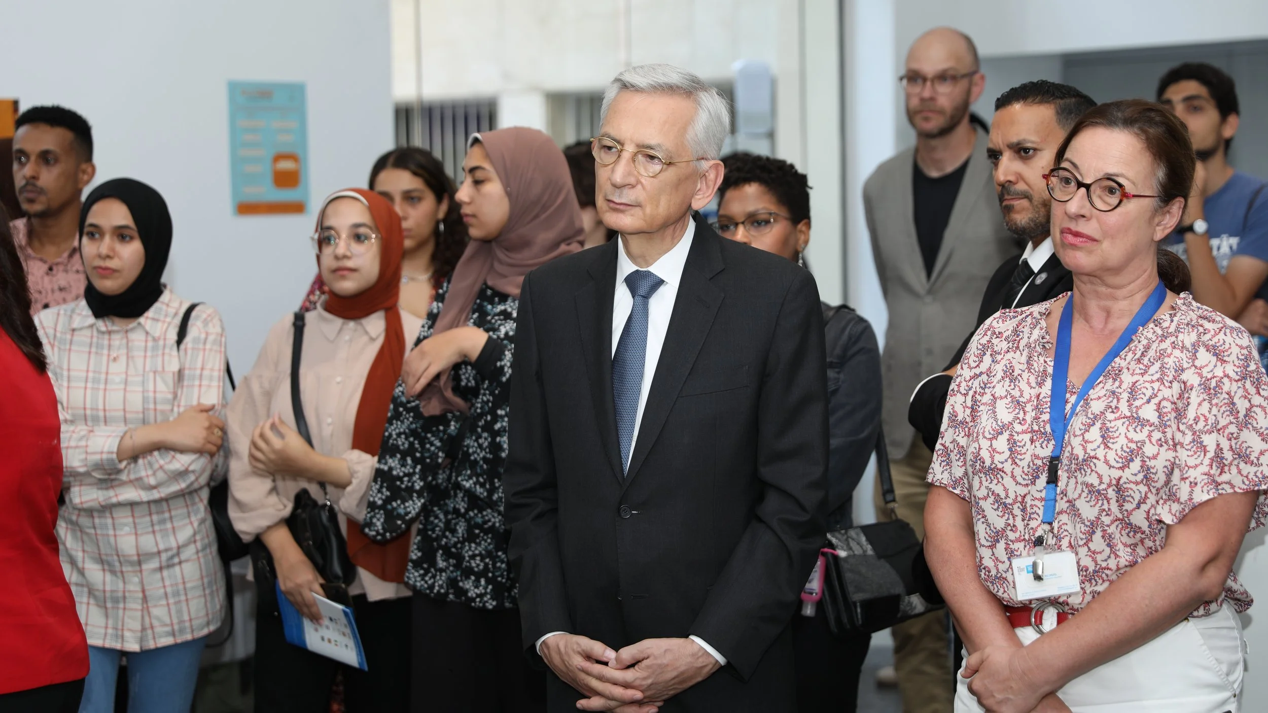 A diverse group of people listening attentively at an indoor event, including an older man in a suit and glasses, a woman with glasses and a floral blouse, and several young adults with varied ethnic backgrounds.
