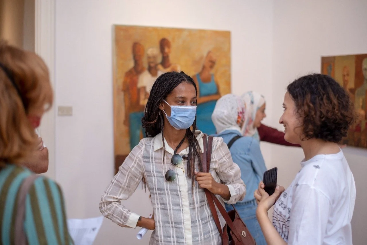 People talking in an art gallery, with artworks on the walls in the background. One woman wearing a face mask is engaged in conversation with another woman holding a phone.