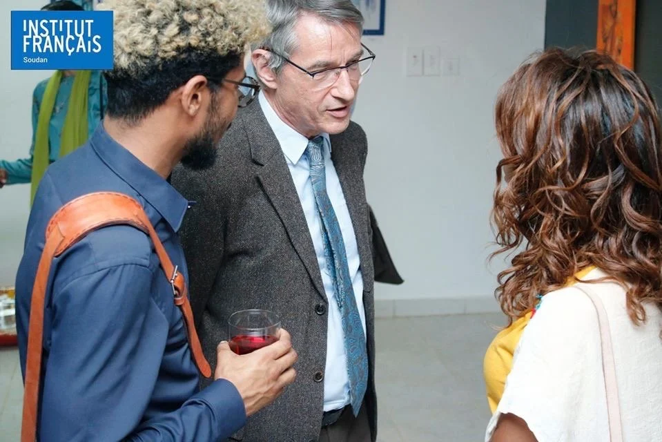 Three people are having a conversation at a social event. One man is holding a glass of red wine, and they are engaged in a discussion. The background shows a sign that says 'Institut Français Soudan'.