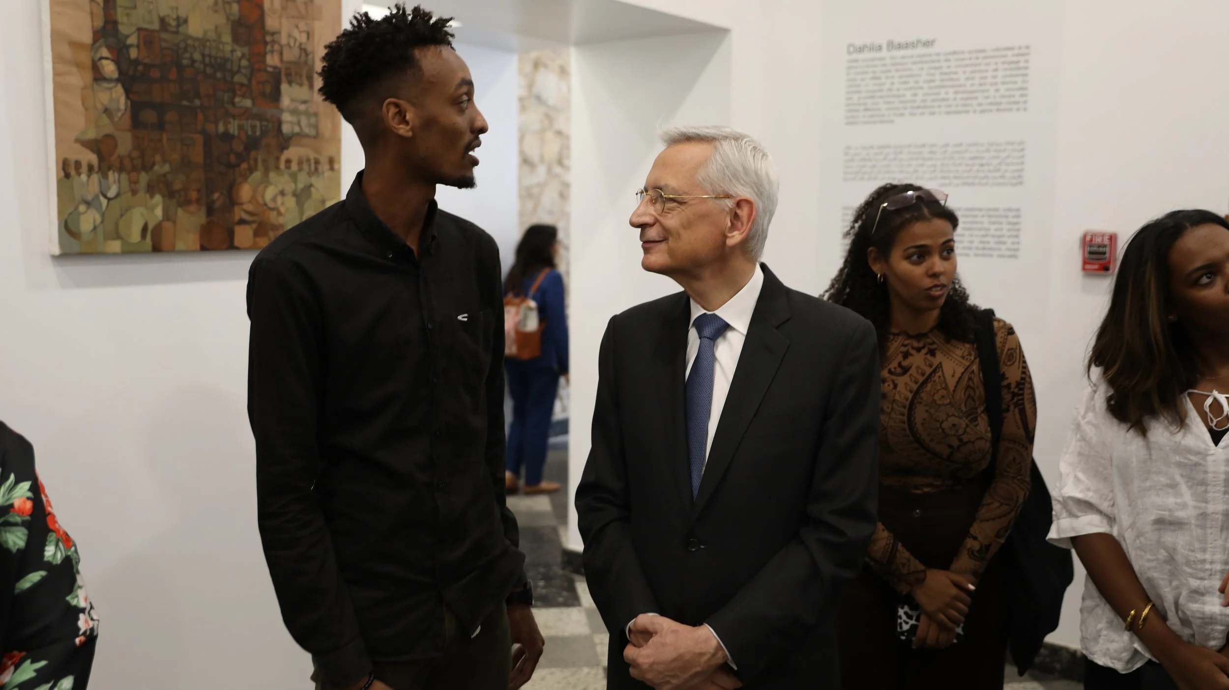 Two men, one young Black man in a black shirt and an older white man in a suit, are talking to each other and smiling at each other in an art gallery. There are other people in the background and abstract artwork on the walls.