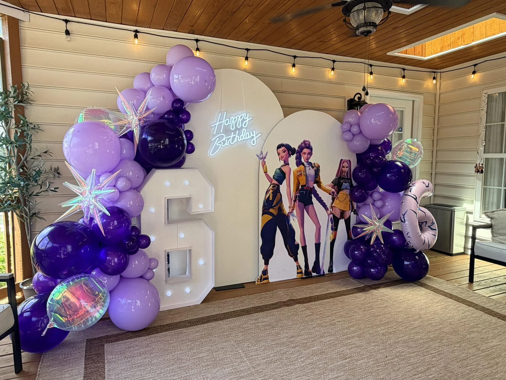 Purple and black balloons surrounding a white backdrop with a photo of five animated women and a illuminated 'Happy Birthday' sign inside a room with wood-paneled ceiling and a window.