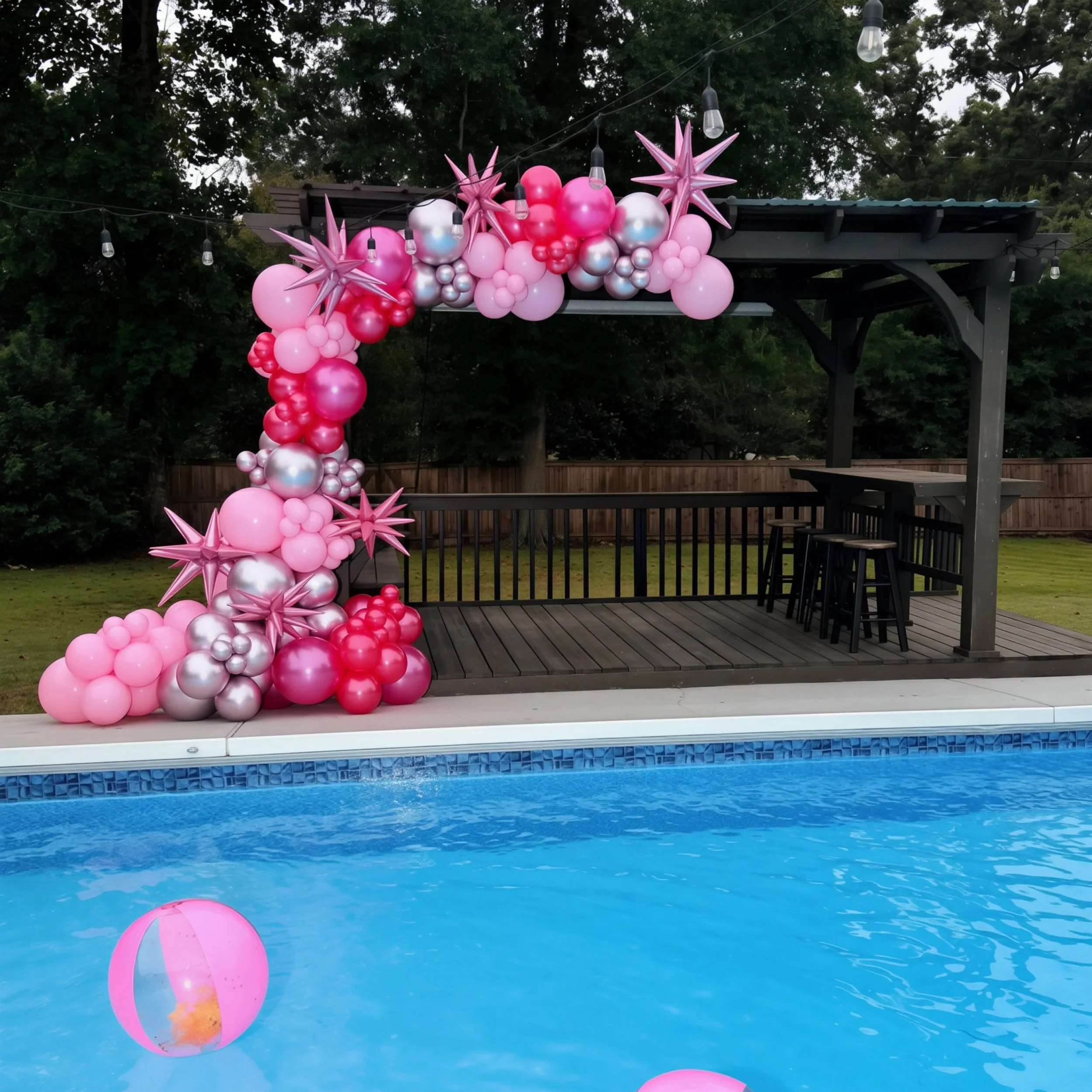 Decorative pink, white, and silver balloons arranged on a deck, with colorful star-shaped balloons and string lights, overlooking a swimming pool with blue water and a pink beach ball floating.