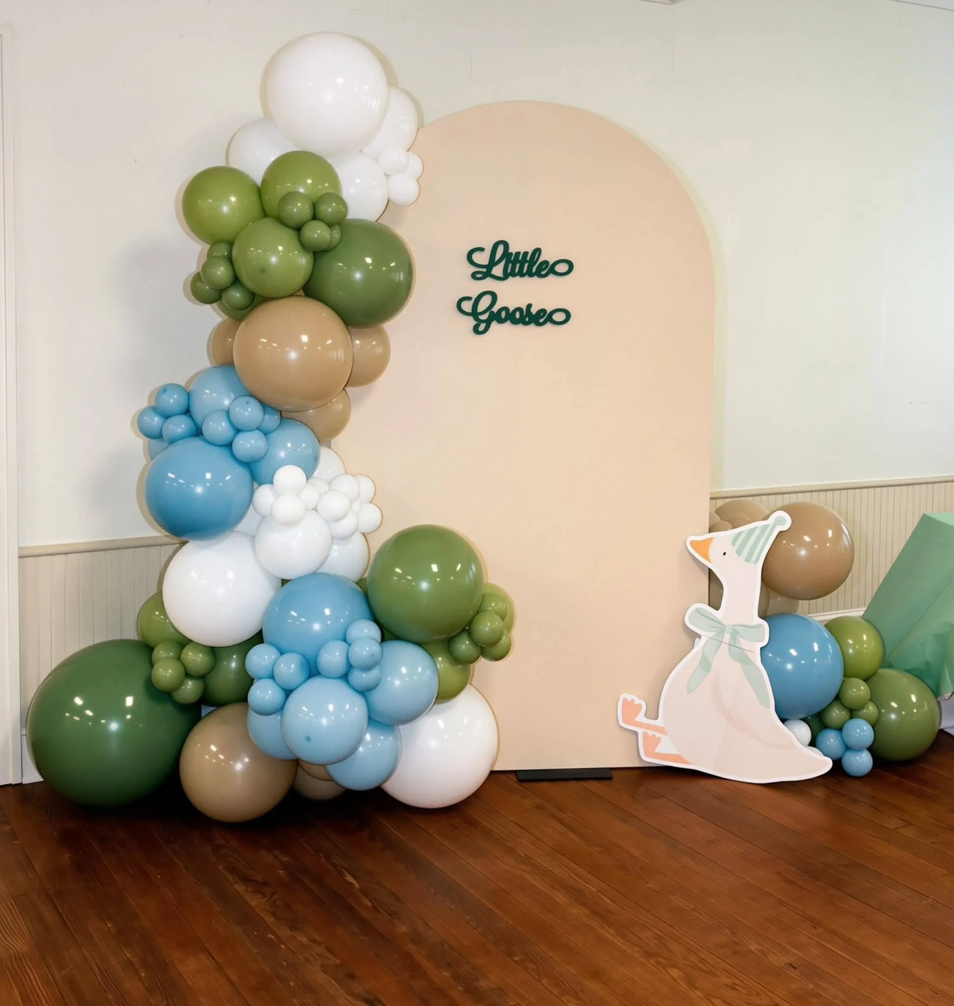 Decorative balloon arrangement with pastel colors including white, beige, light blue, and green, alongside a paper cutout of a baby wearing a party hat, for a baby shower celebration.