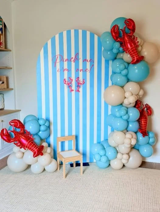 A seaside-themed backdrop with blue and white vertical stripes, decorated with red lobster balloons and clusters of blue and white balloons. The backdrop has the phrase "Hug me, I'm one!" written in pink. A small wooden chair is in front of the display.