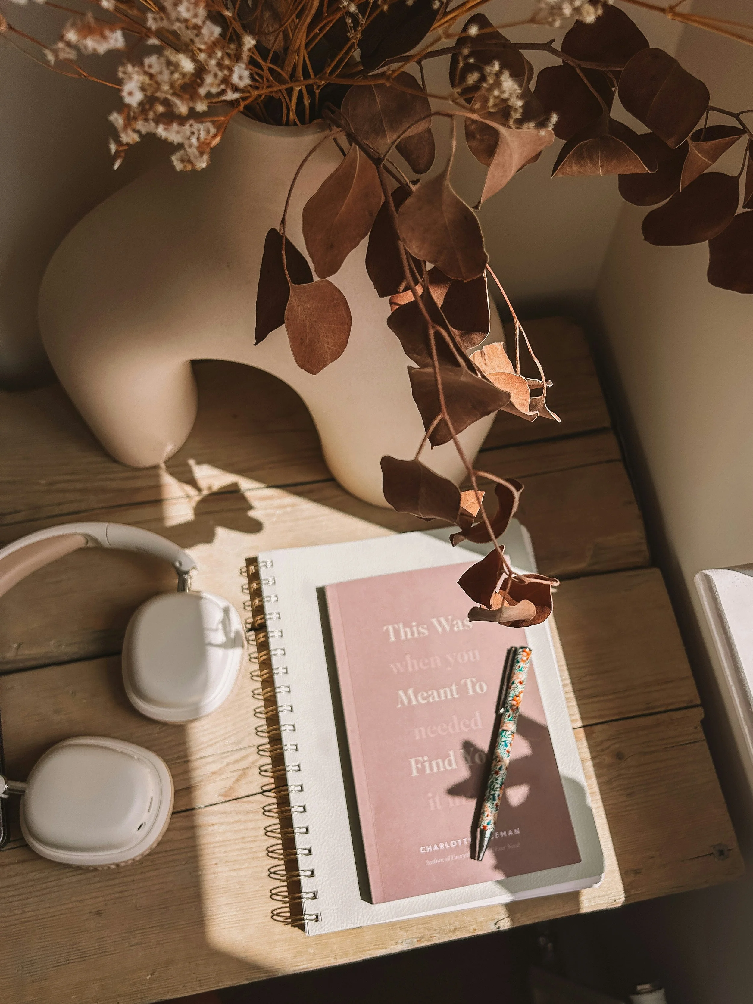 Warmly lit wooden desk with white vases of dried eucalyptus, a pink devotional book, floral pen, notebook, and earbuds.