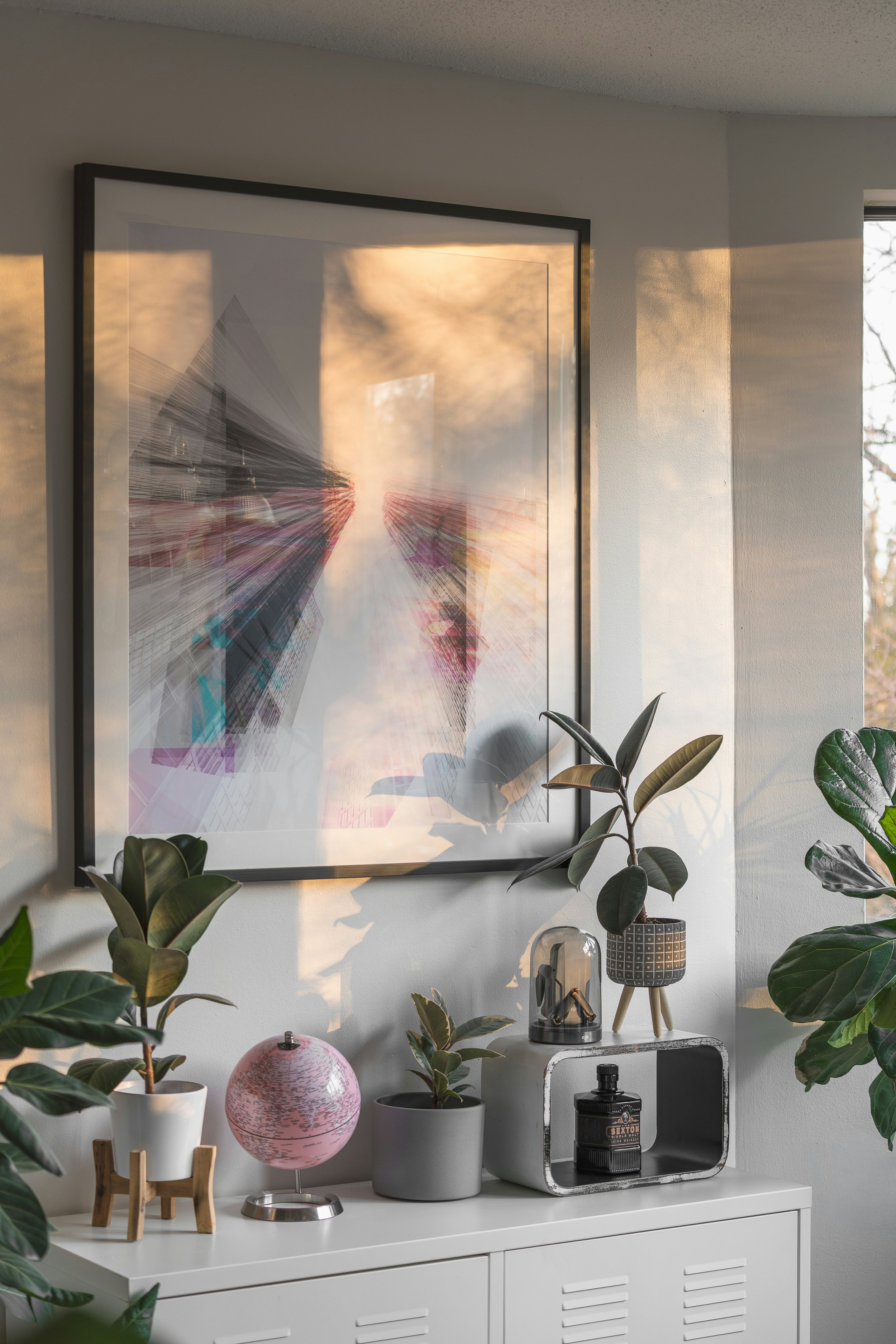 Sunlit modern living room console with framed abstract artwork, potted houseplants, pink globe, decorative sculpture, and whiskey bottle.