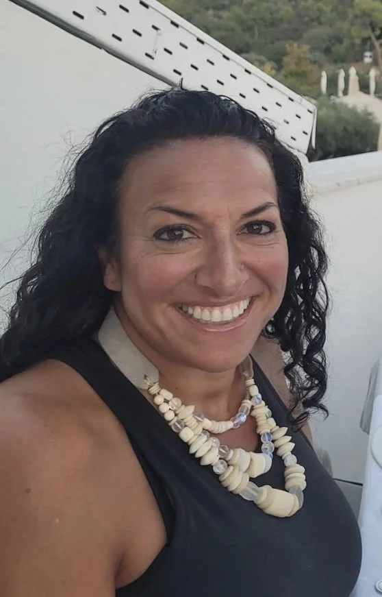 Smiling woman with curly dark hair wearing a black top and layered shell necklace outdoors